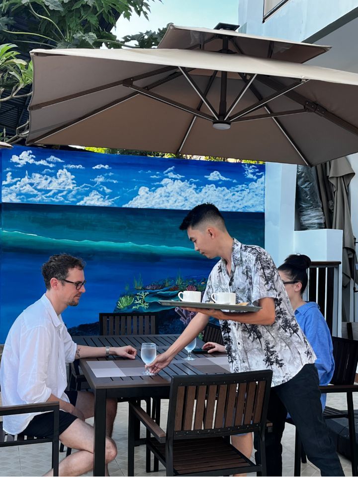 A man is carrying a tray of food to a group of people sitting at a table under an umbrella.