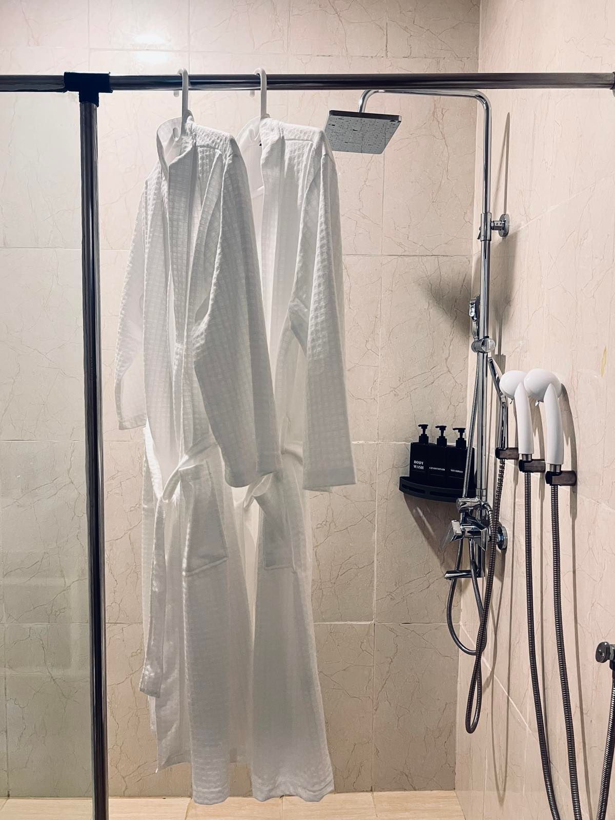 Two white robes are hanging in a shower with a shower head.