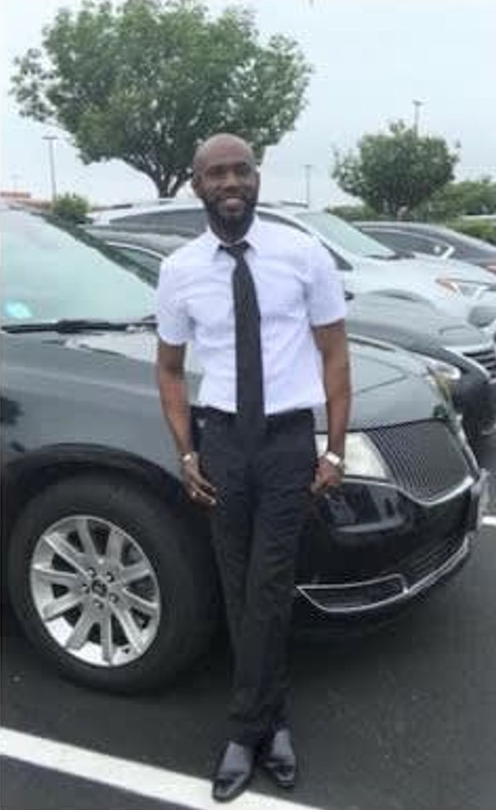 A man in a suit and tie is standing in front of a black car in a parking lot