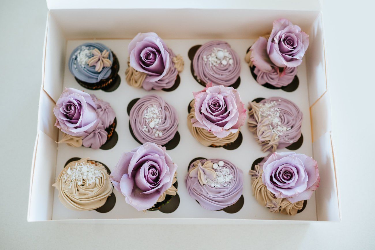 A box of cupcakes with purple frosting and purple roses.