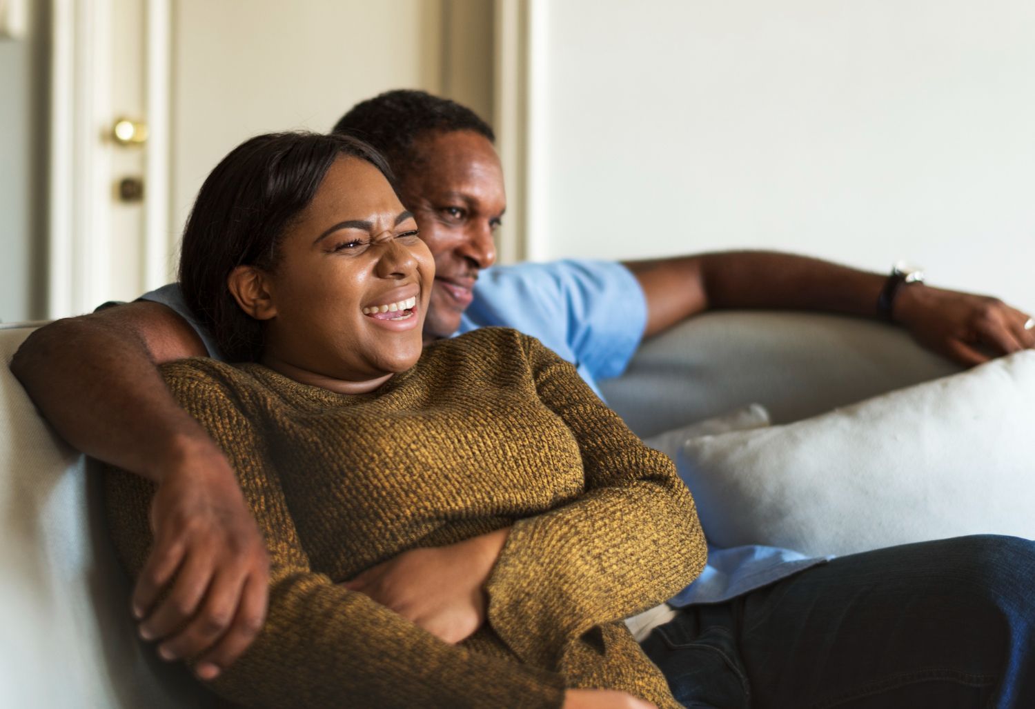 Couple on a couch smiling and embracing. Woman in a brown sweater, man in a blue shirt. Interior setting.