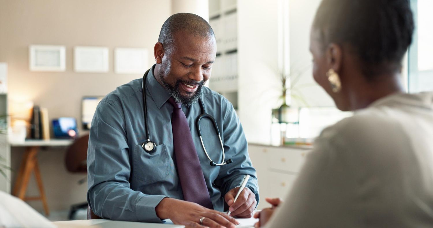 Doctor in blue shirt with stethoscope, writing at desk while talking with a patient.