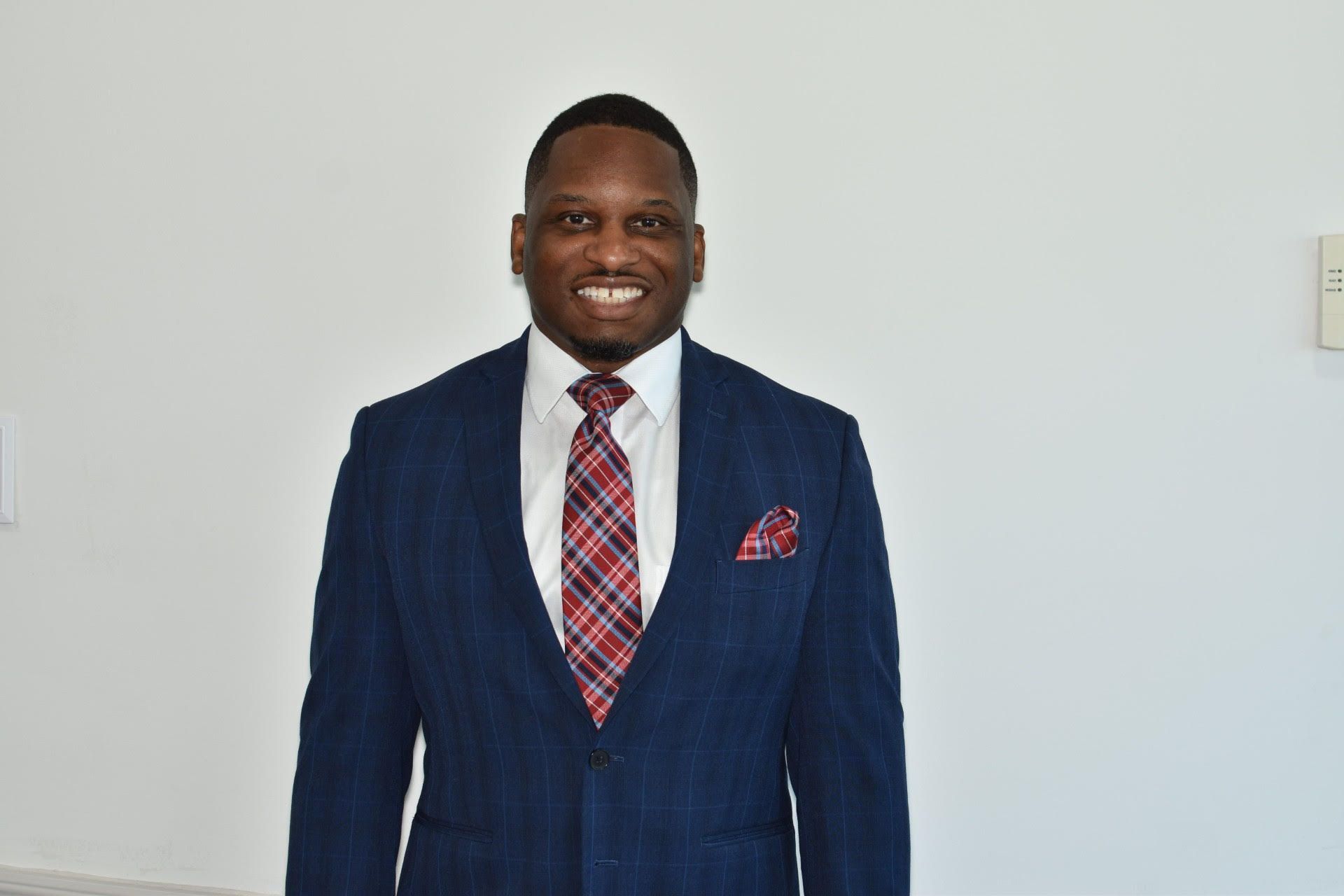 Man in blue suit, red tie, and pocket square smiles in front of a white wall.