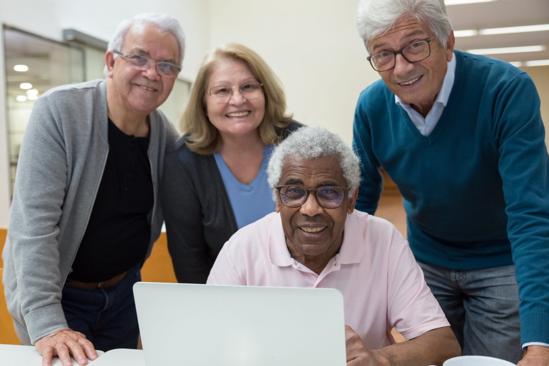 Group of seniors around a laptop, smiling. Inside a building, wearing casual clothes.