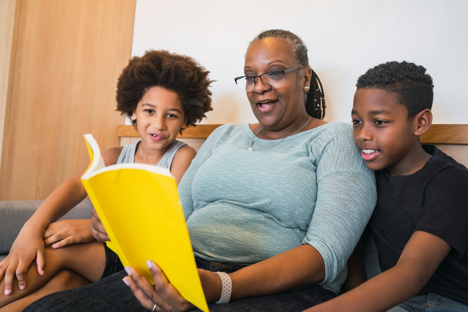 Woman reading aloud to two children from a yellow book; they are all smiling indoors.