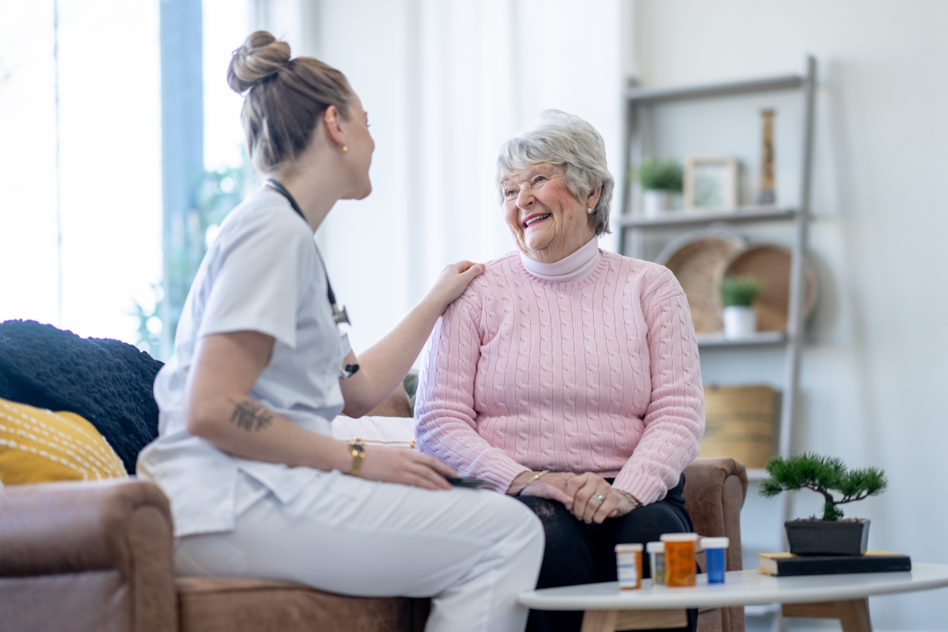 Nurse comforts elderly patient, hand on her shoulder. They smile, seated in a living room; pills on a table.