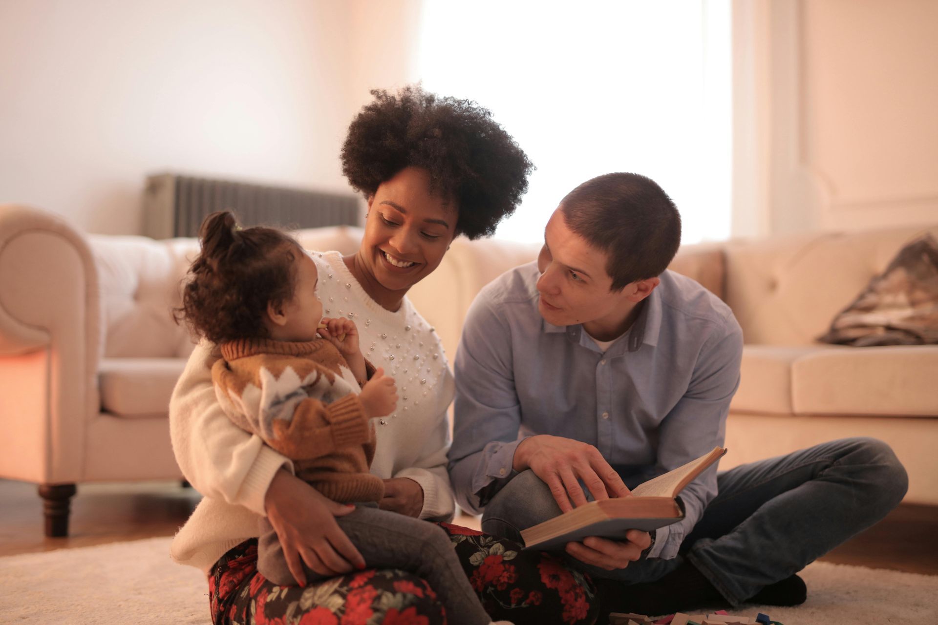 Family reading together on floor. Woman holds child, man reads book. Living room setting.