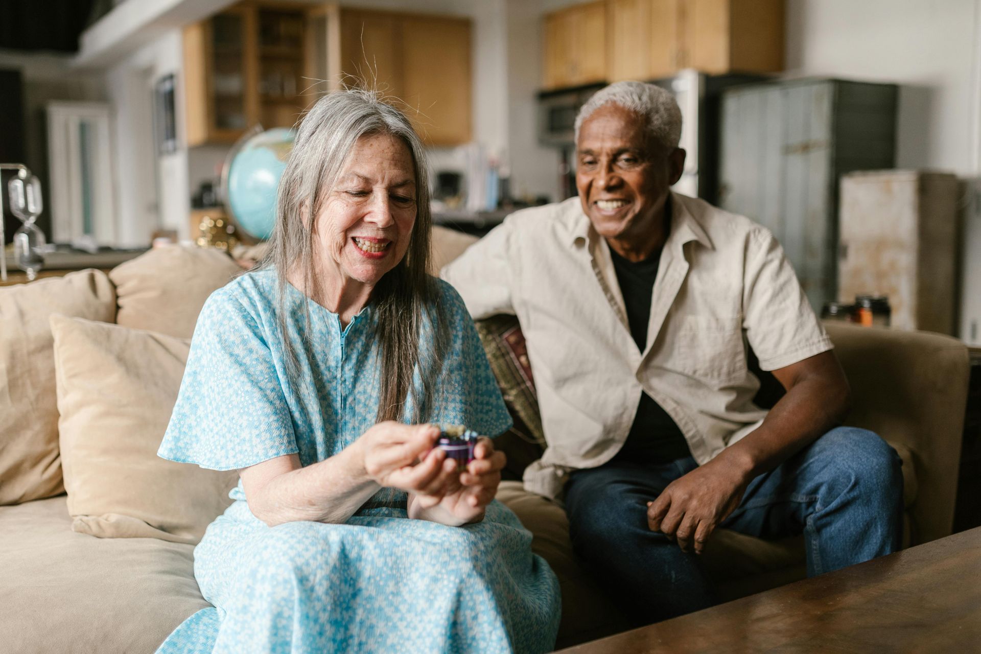 An older person smiles while holding a purple object; another person smiles, both seated on a couch indoors.