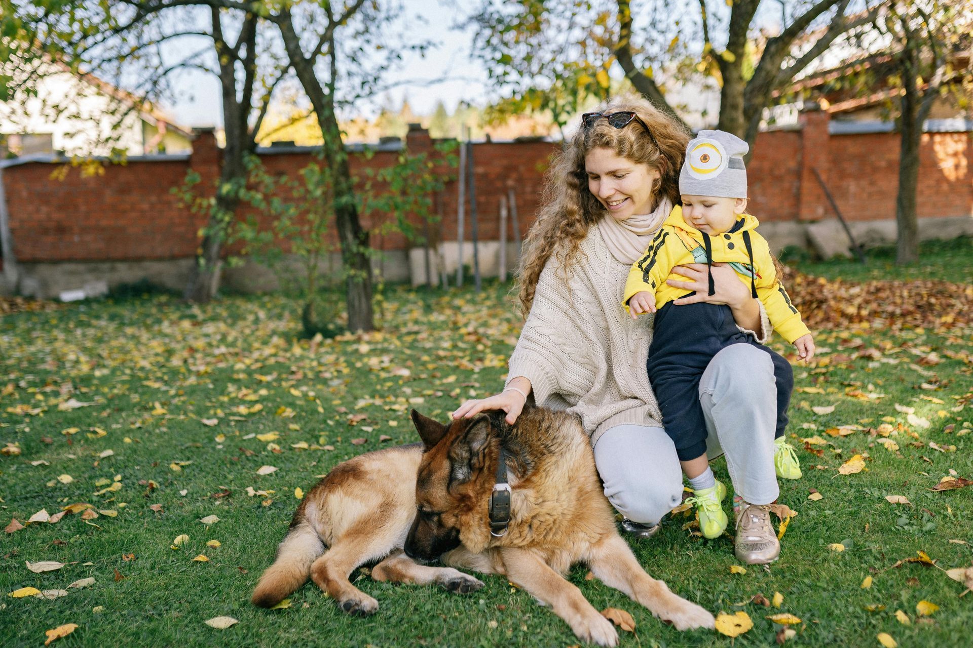 Woman petting dog while holding a baby on a green lawn with fallen leaves.