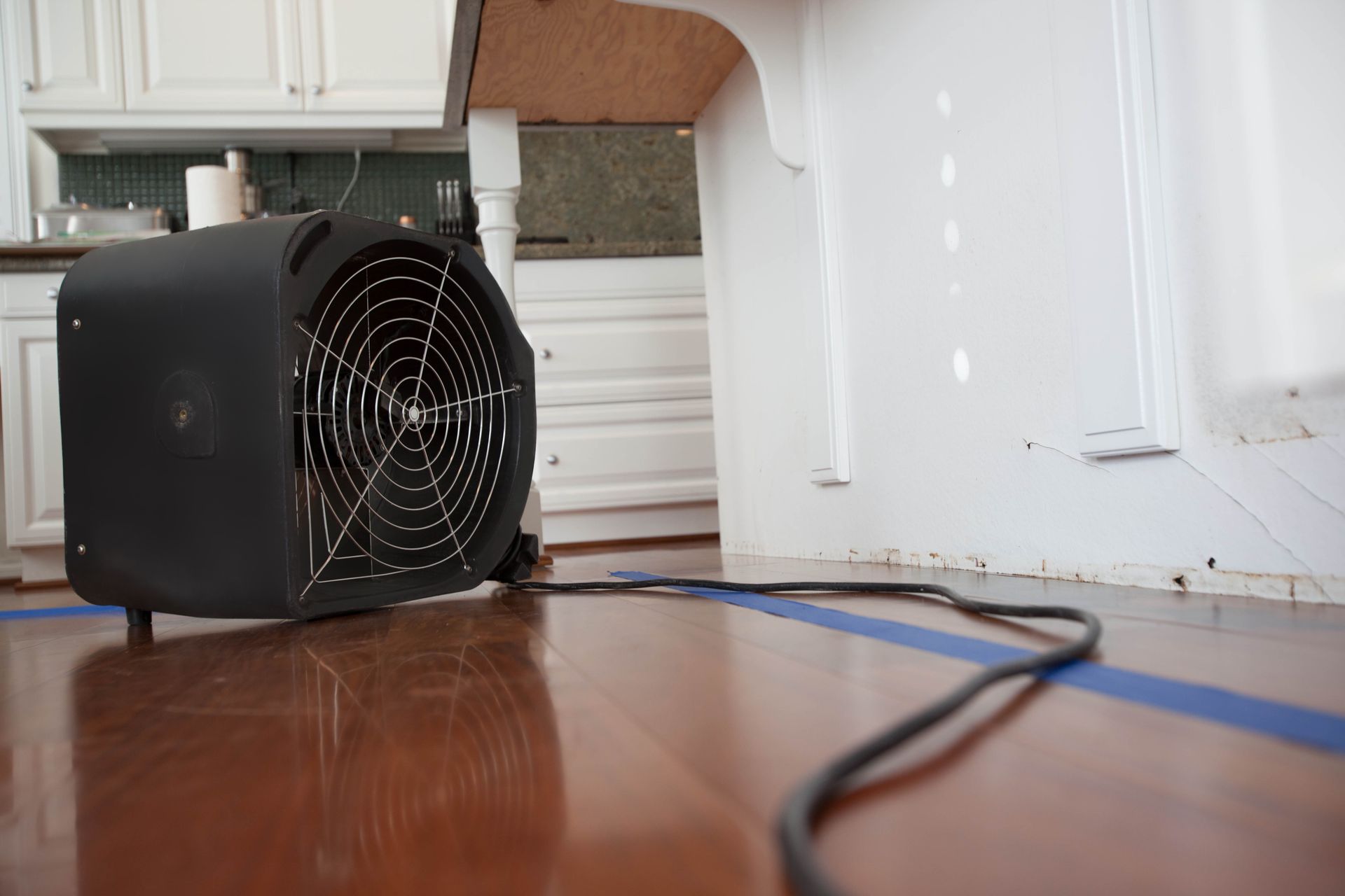 A black fan is sitting on a wooden floor in a kitchen.