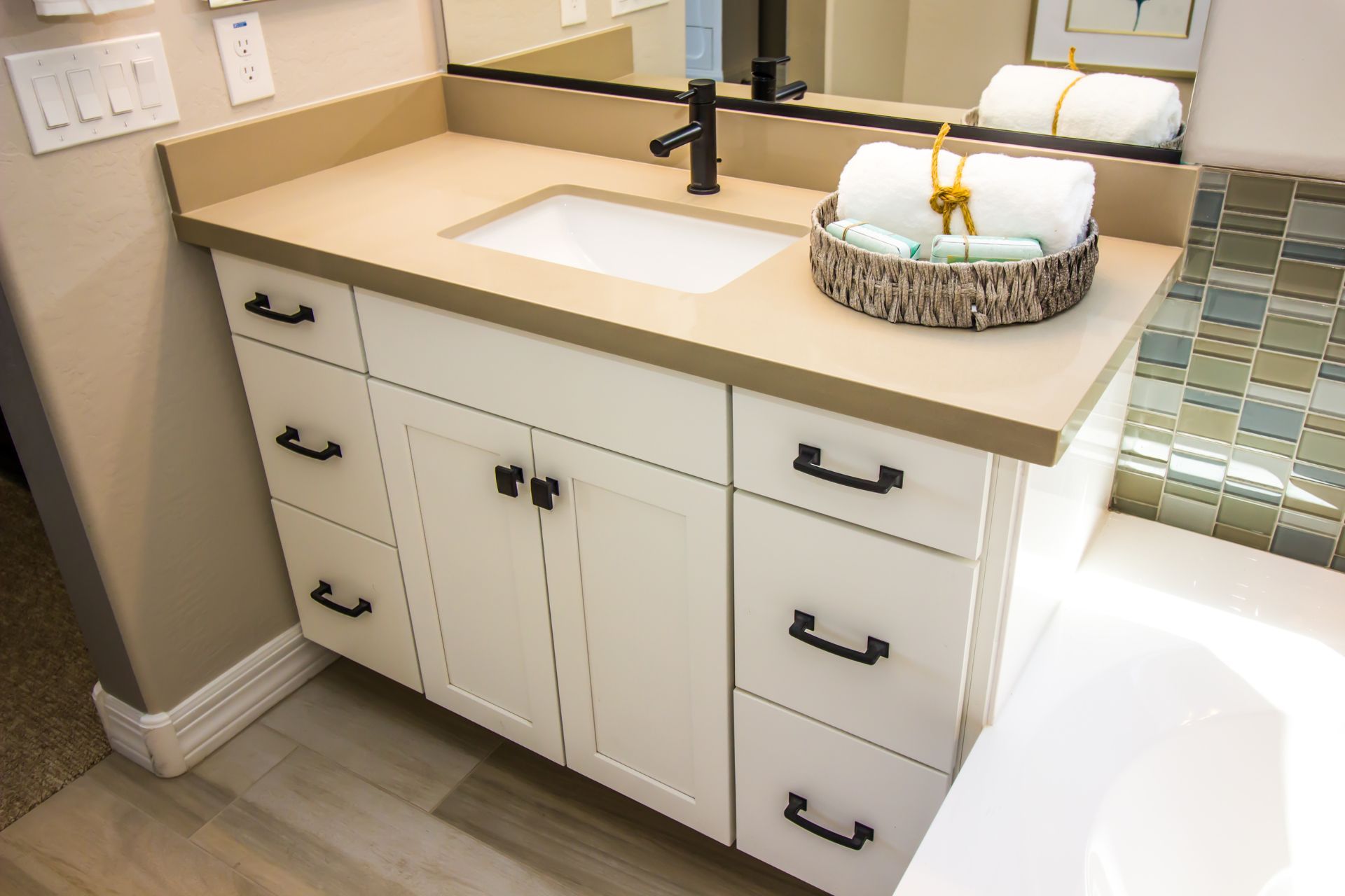 White bathroom vanity with black fixtures, tan countertop, and wicker basket with towels.