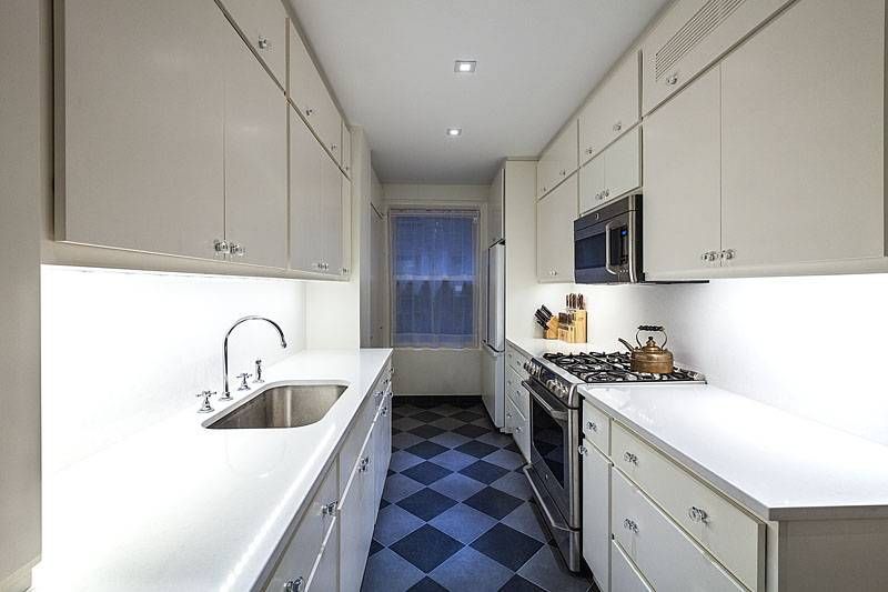 Long, narrow, white kitchen with checkerboard floor. Stainless steel appliances, overhead cabinets, sink, and window.