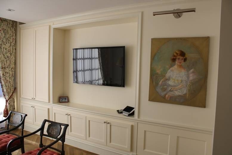 Cream-colored wall unit with a TV, painting, and storage cabinets, next to curtains and two chairs.