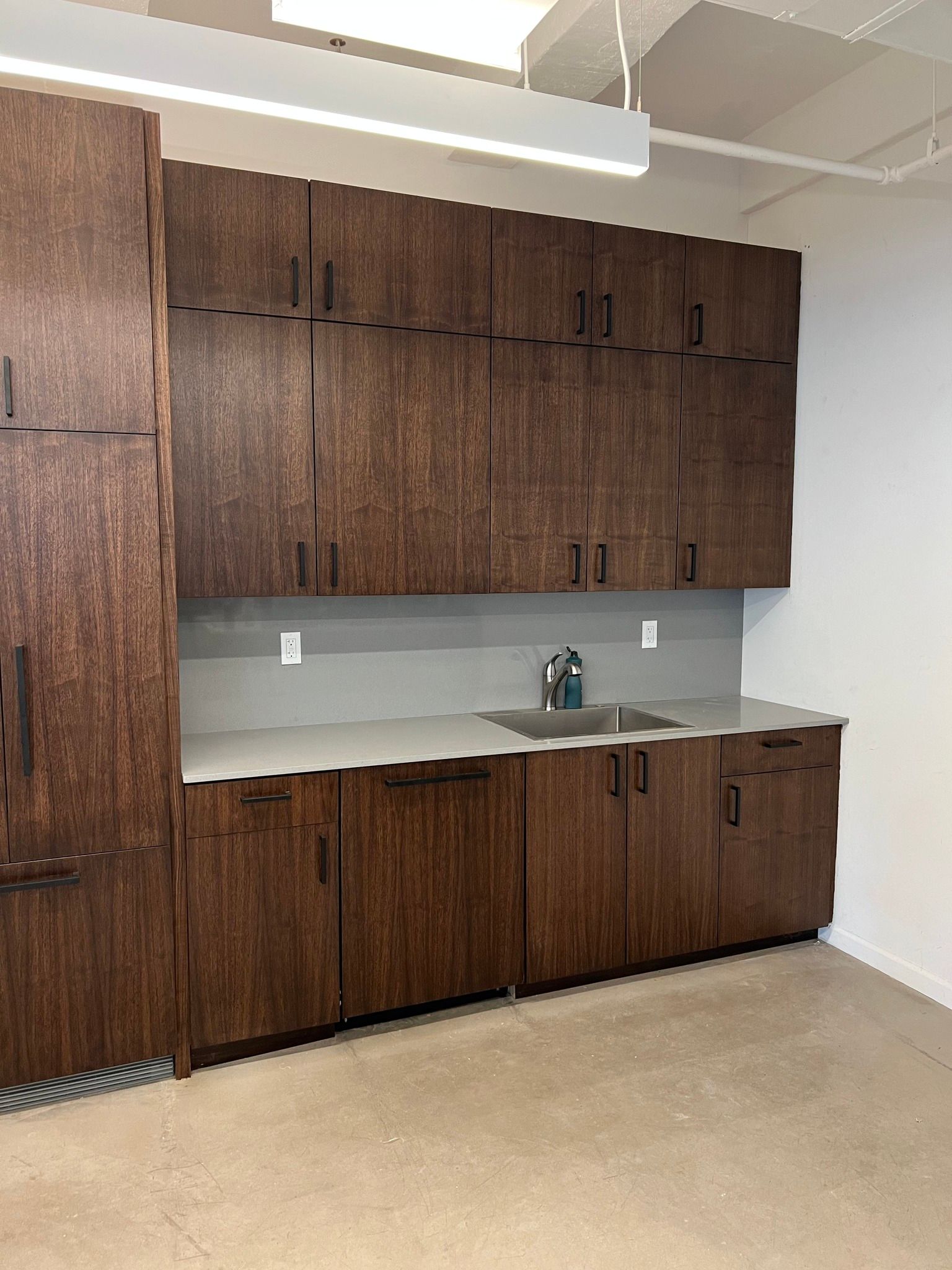 Dark wood cabinetry with a sink, counter, and upper cabinets in a modern office space.