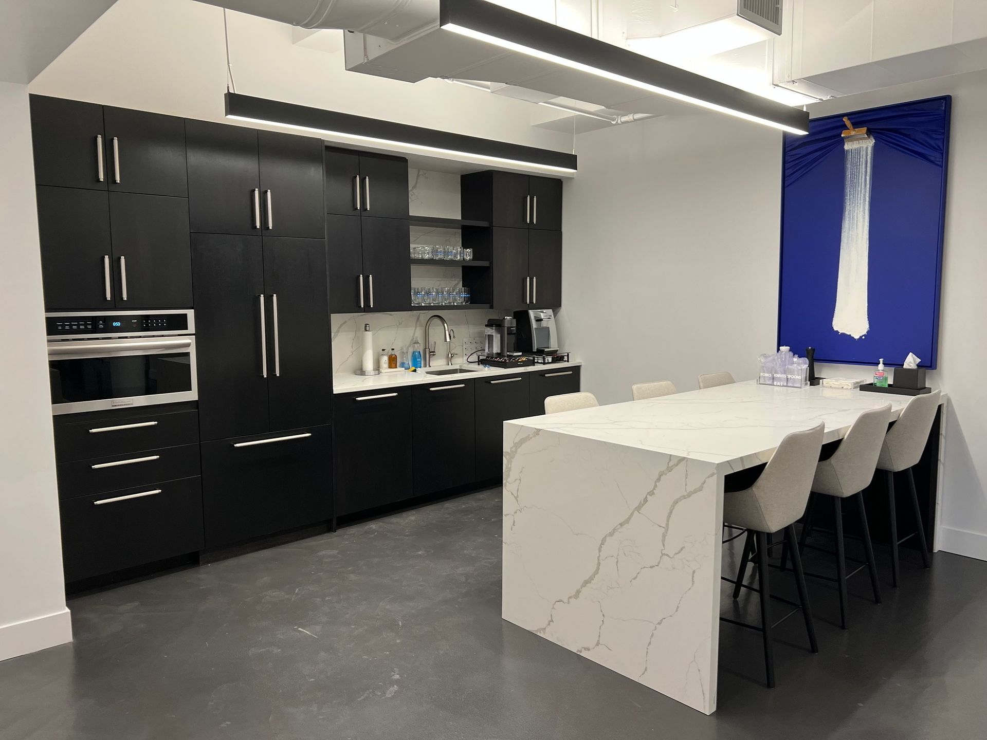 Modern office kitchen with black cabinets, white countertops, and a marble-look island.