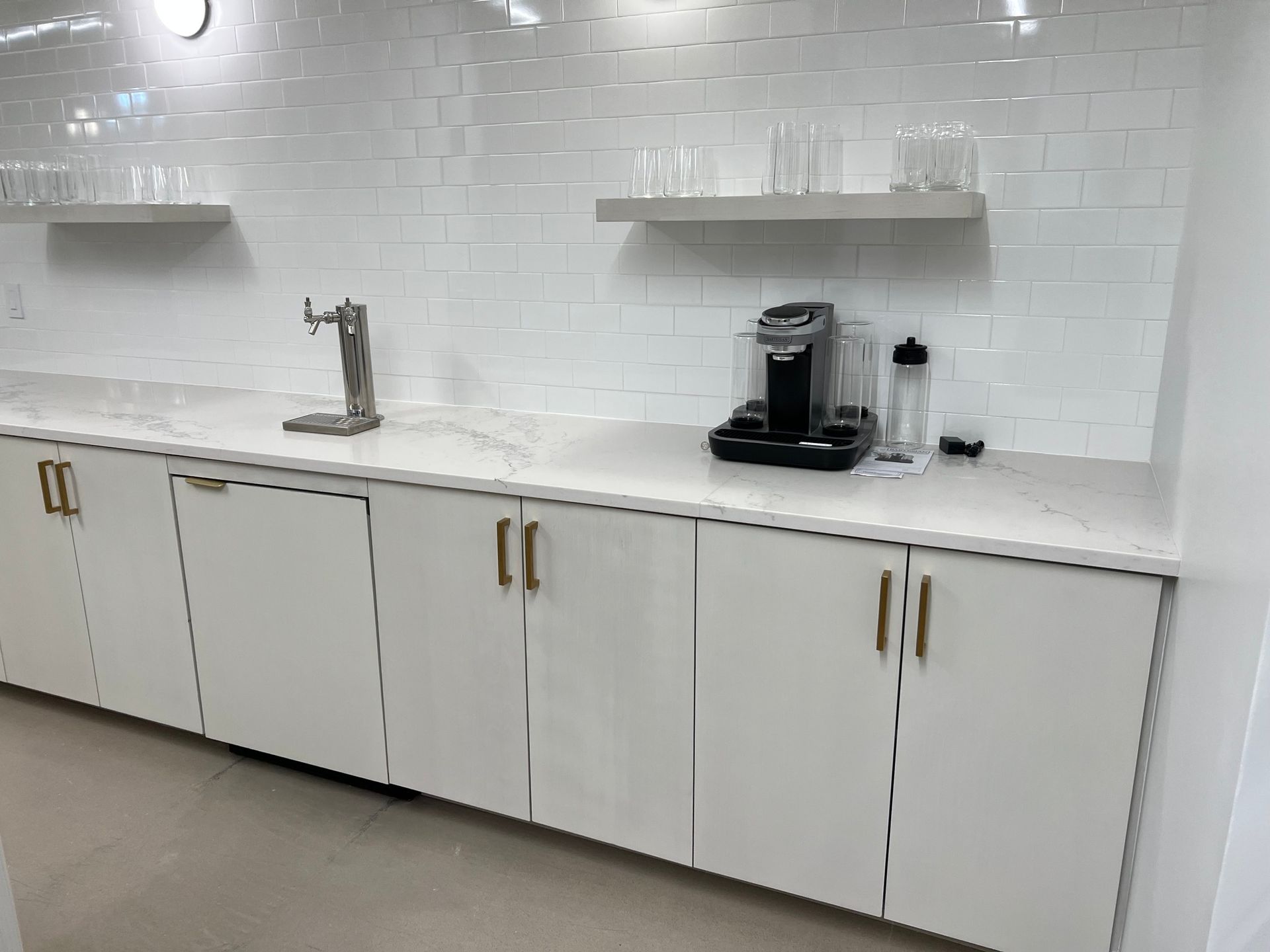 White kitchenette with cabinets, countertop, water dispenser, Keurig, and shelves holding glasses.