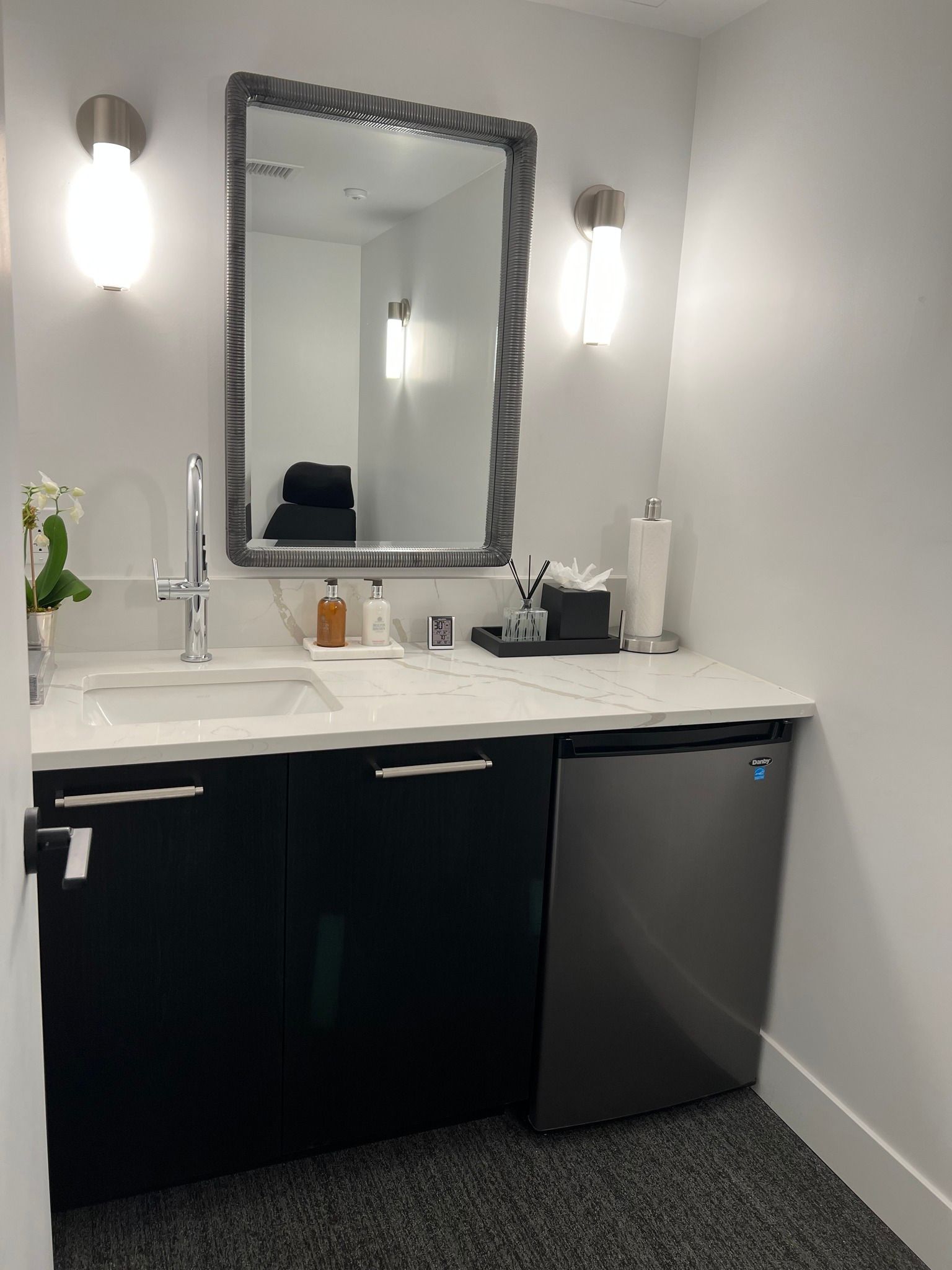 Small bathroom with a sink, mirror, and mini-fridge. Black cabinets, white countertop, and gray carpet.
