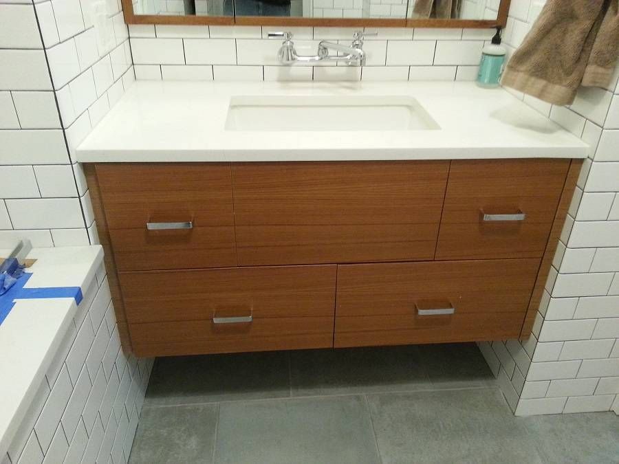 Floating wooden bathroom vanity with white countertop and sink, mounted on tiled wall.