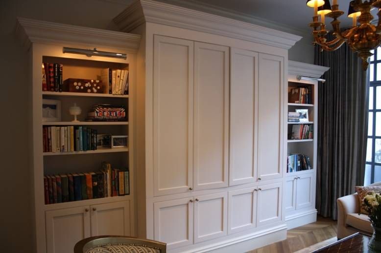 Built-in white bookcase with cabinets, bookshelves, and overhead lighting in a room with a window and ornate chandelier.