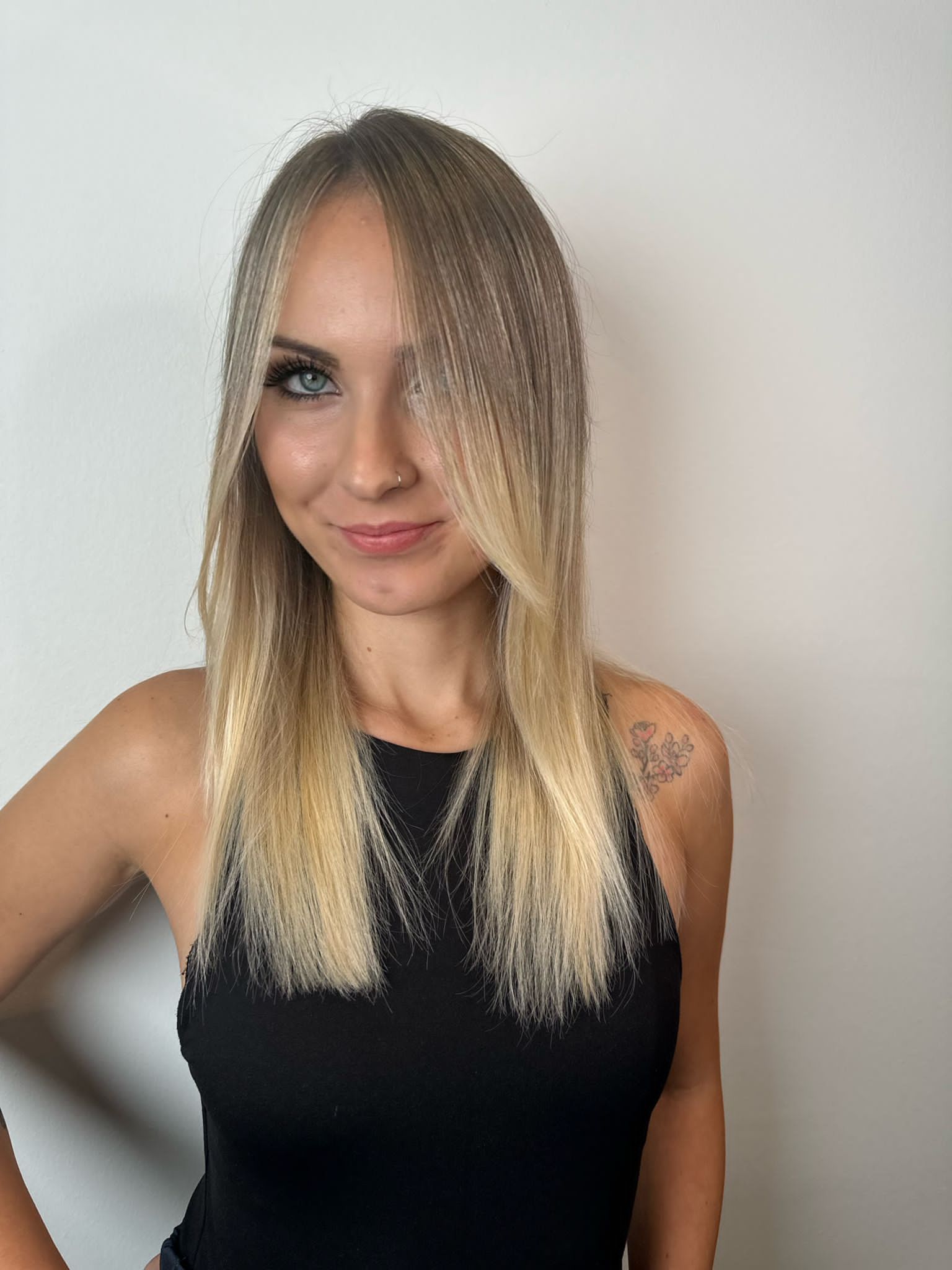 Una persona con lunghi capelli biondi ondulati e un piercing al naso, che indossa una maglietta nera su uno sfondo bianco uniforme.