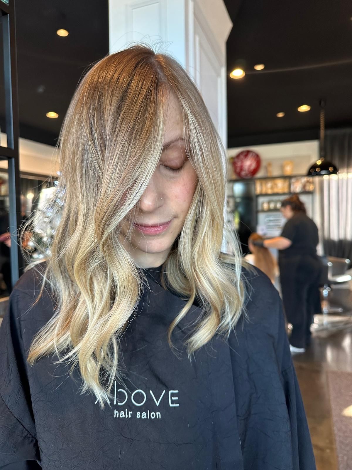 Una persona con i capelli mossi e biondi con mèches, in un salone di parrucchiere, indossa un mantello nero da parrucchiera.