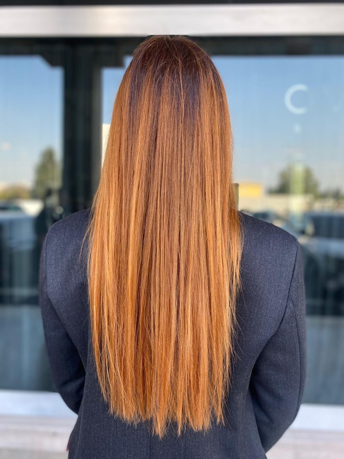 Una persona con lunghi capelli lisci color rame, vista di spalle, indossa un blazer blu scuro ed è in piedi davanti a una vetrata.