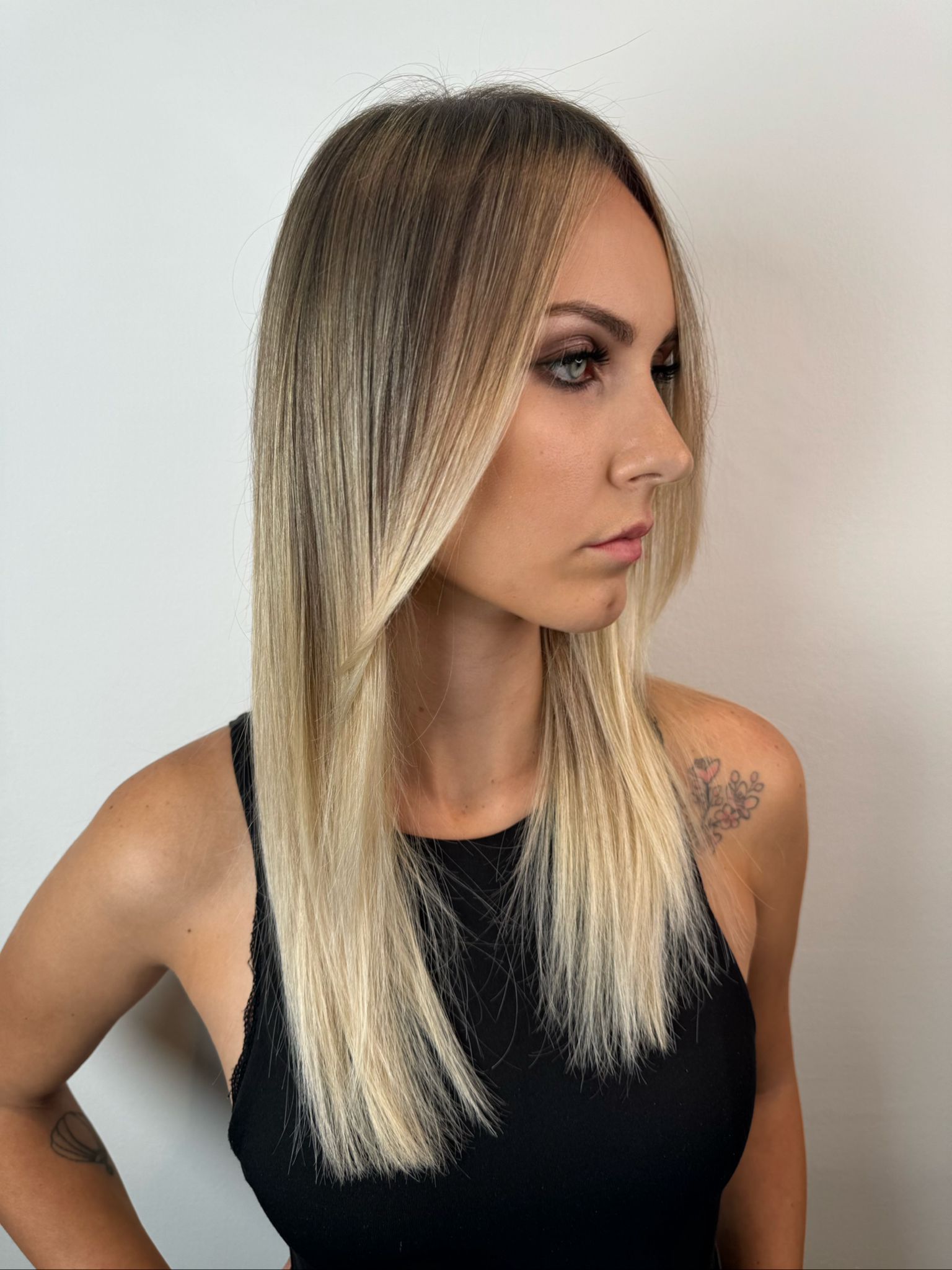 Una persona con lunghi capelli biondi ondulati con radici scure, che indossa una canottiera nera su uno sfondo bianco uniforme.