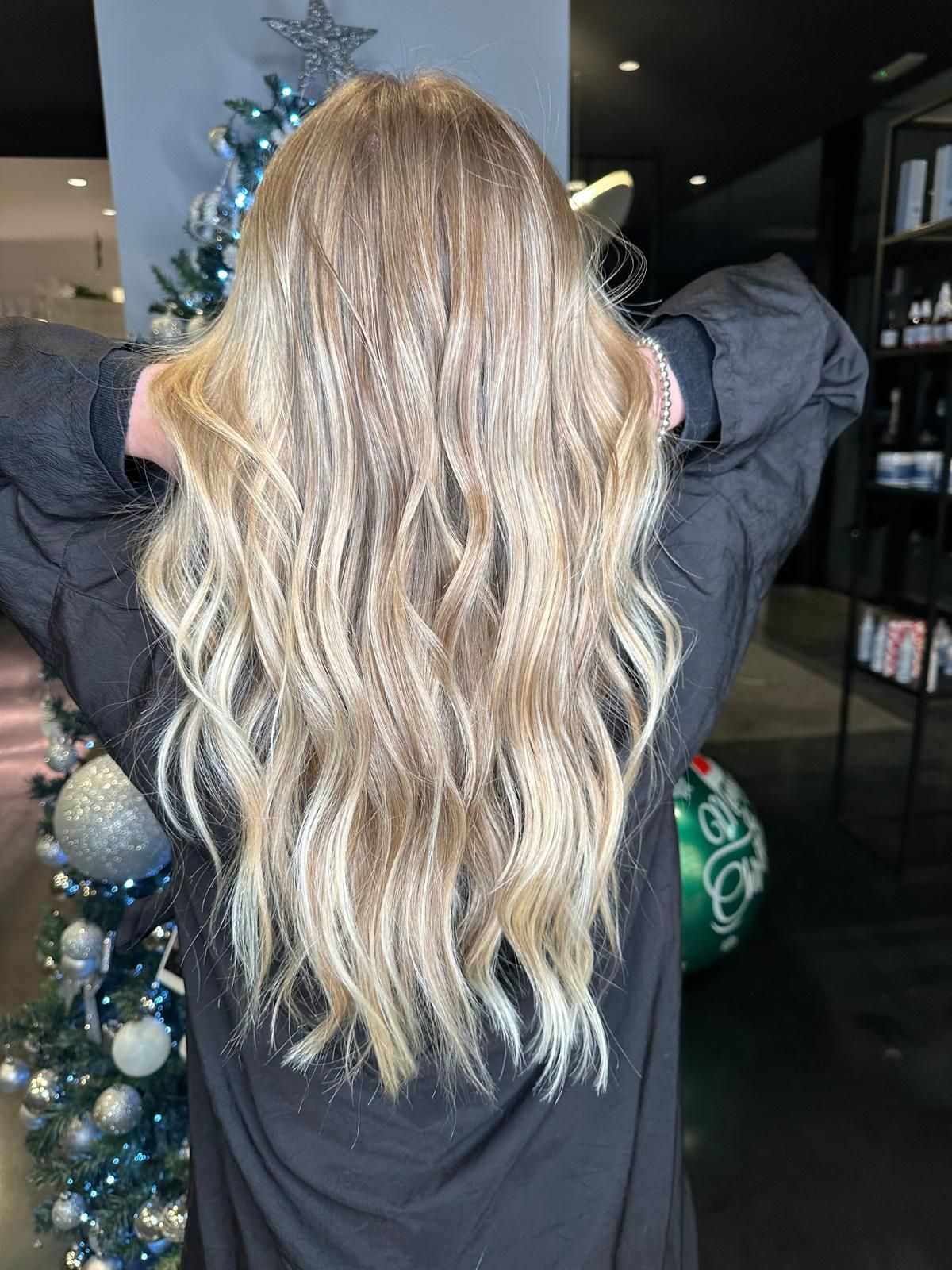 Lunghi capelli biondi, mossi e con mèches, visti di spalle in un salone di bellezza, con decorazioni natalizie sullo sfondo.