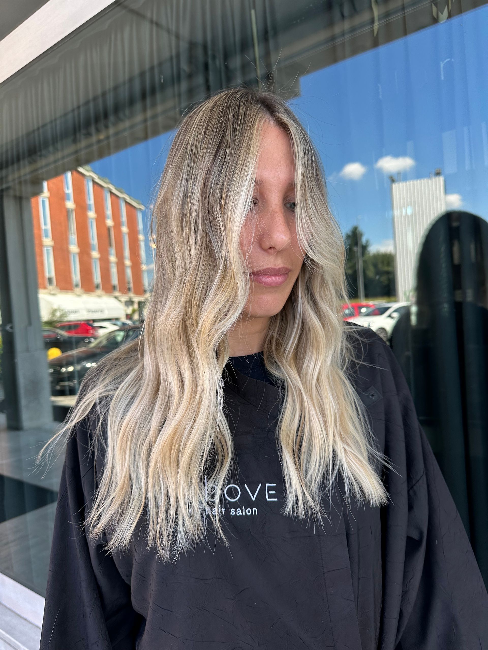 Una persona con lunghi capelli biondi, mossi e dalla texture particolare, è in piedi davanti a una finestra che riflette un edificio cittadino e il cielo azzurro.