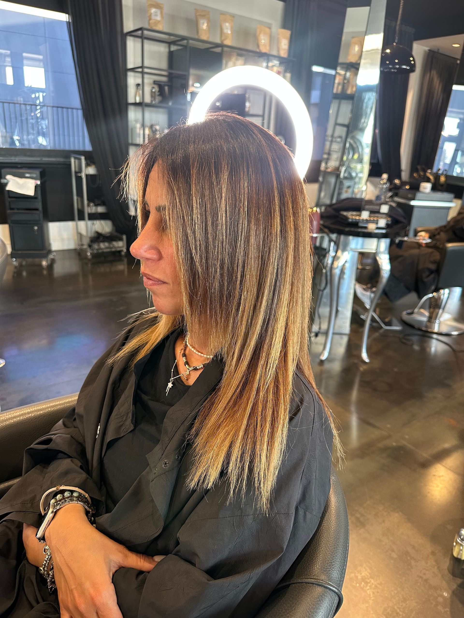 In un salone da parrucchiere, una persona è seduta su una poltrona con i capelli bagnati, lunghi fino alle spalle e con delle mèches, e indossa un mantello nero.