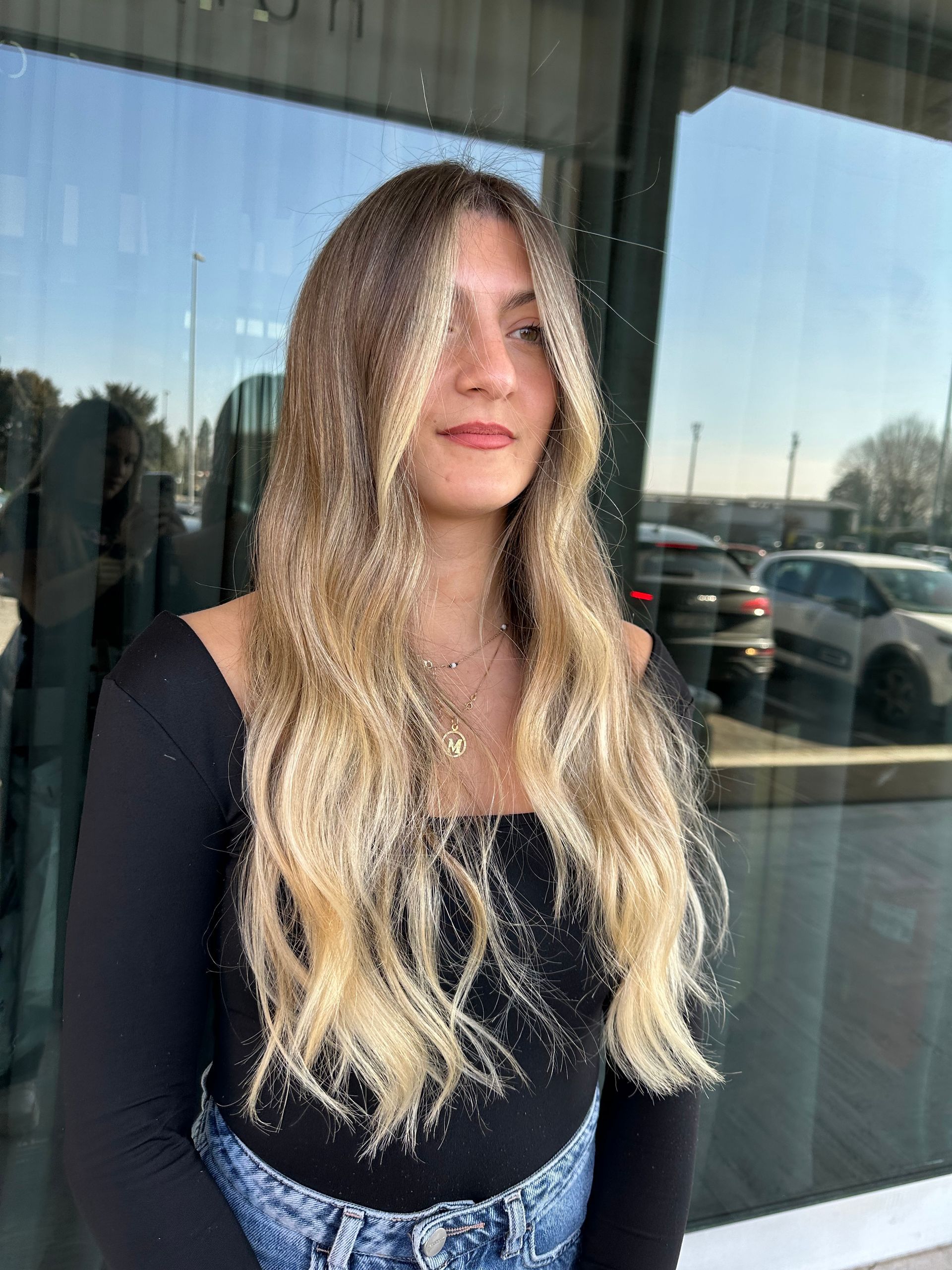 Una persona con lunghi capelli biondi ondulati e mèches, che indossa una maglietta nera, è in piedi all'esterno davanti a un edificio di vetro.