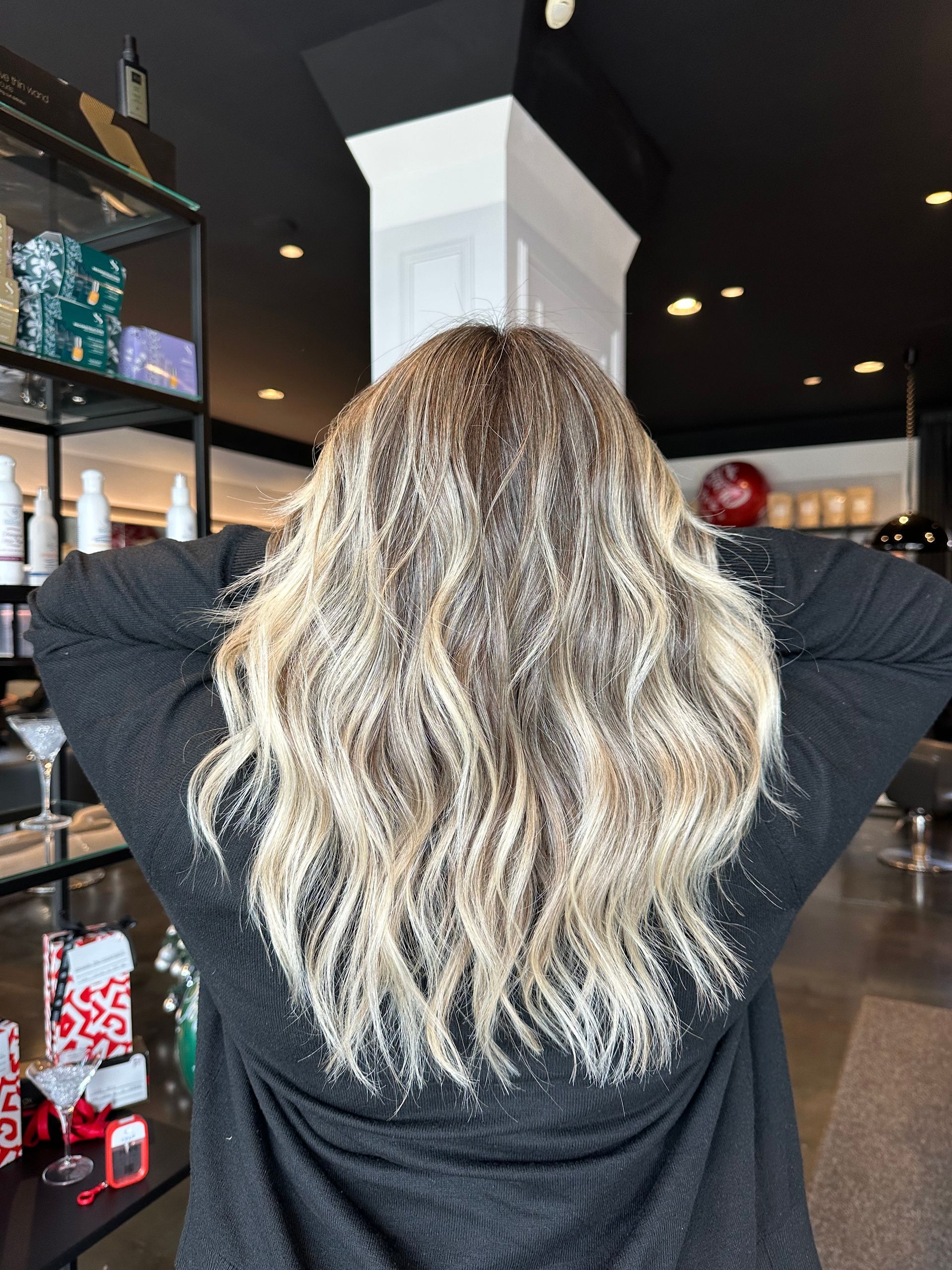 Una persona con capelli biondi, mossi e con mèches, lunghi fino alle spalle, vista da dietro, in piedi in un salone ben illuminato.