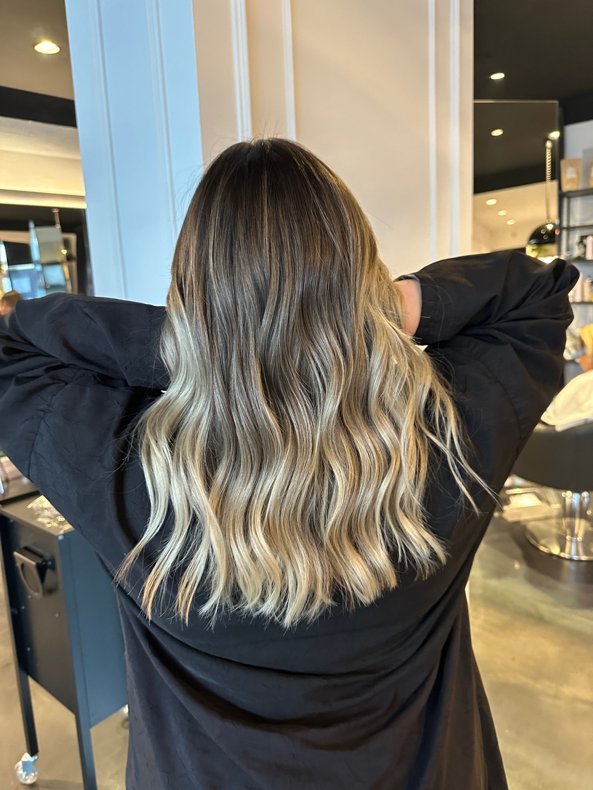 Una persona con capelli mossi di media lunghezza, con radici castano scuro che sfumano in mèches bionde, in un salone di bellezza.