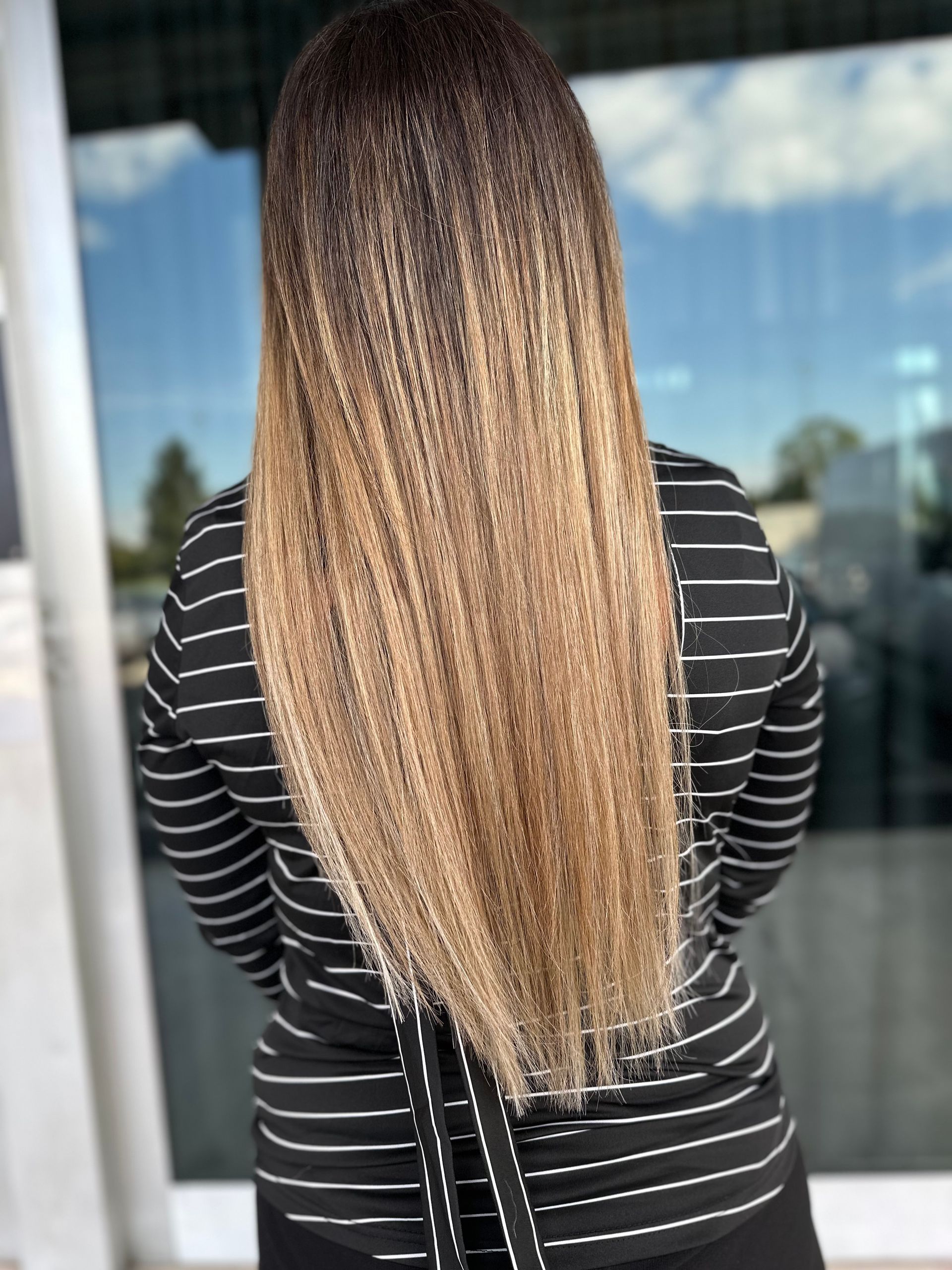 Una persona con lunghi capelli mossi, di colore che sfuma dal scuro al biondo, è in piedi di spalle alla telecamera, con indosso una camicia a righe.