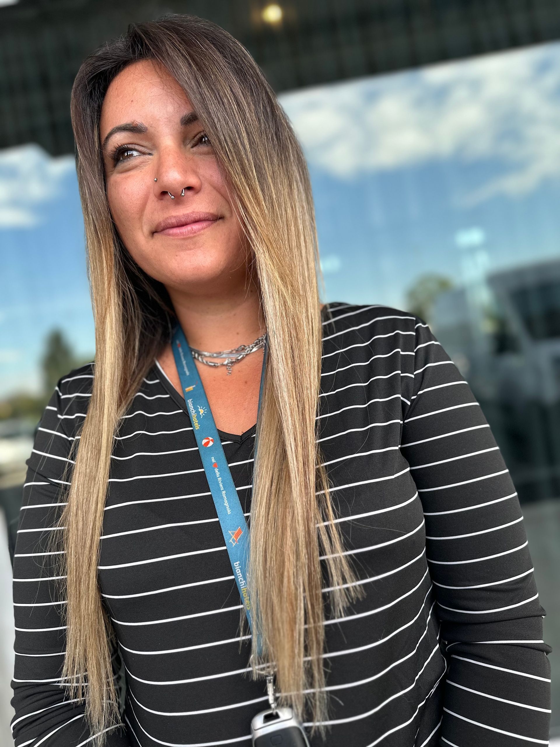 Una persona con lunghi capelli biondi bicolore, che indossa una maglietta a righe bianche e nere e un cordino blu, all'aperto.