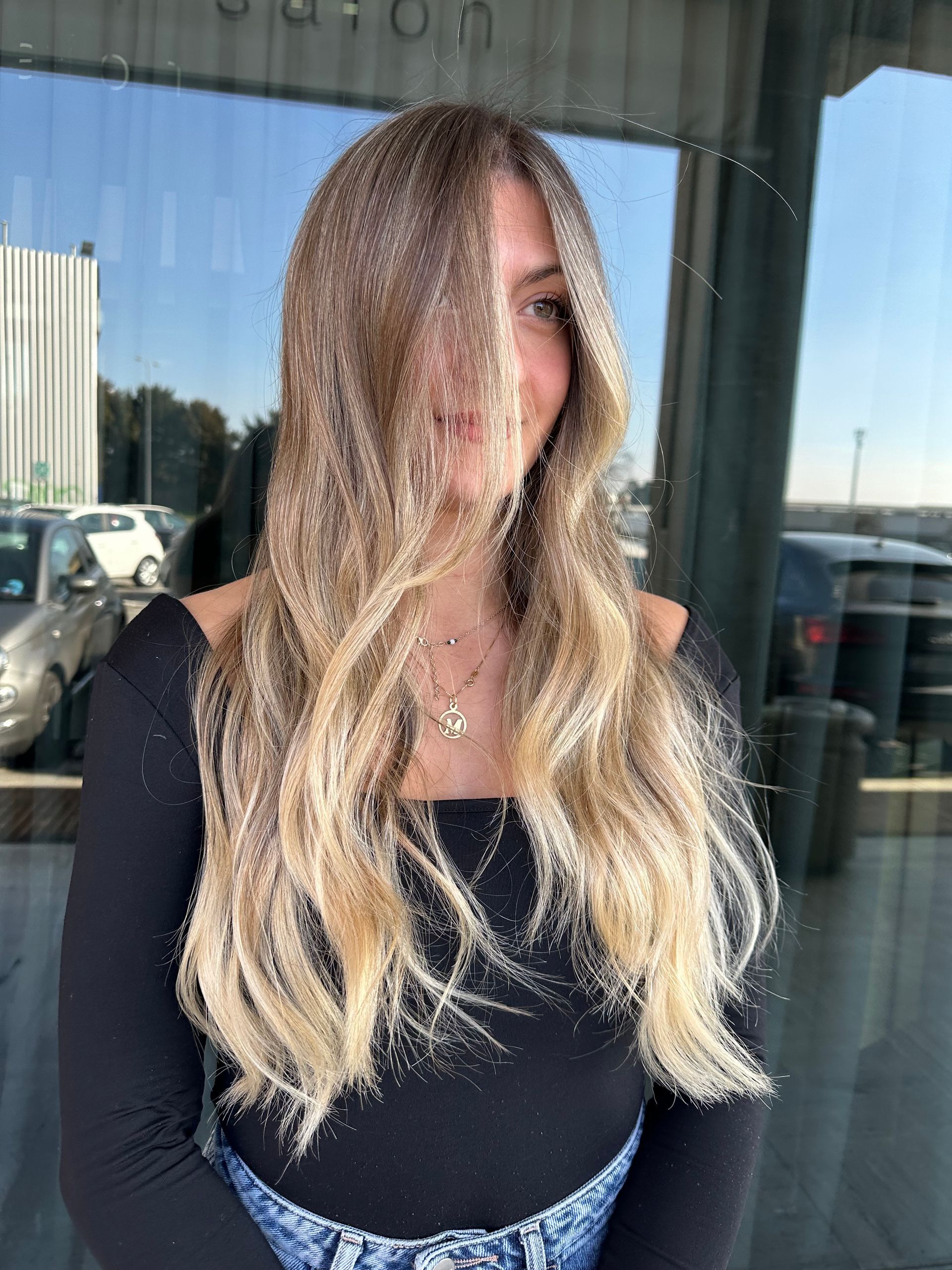 In una giornata di sole, una persona con lunghi capelli biondi ondulati e balayage posa davanti alla vetrina di un negozio.