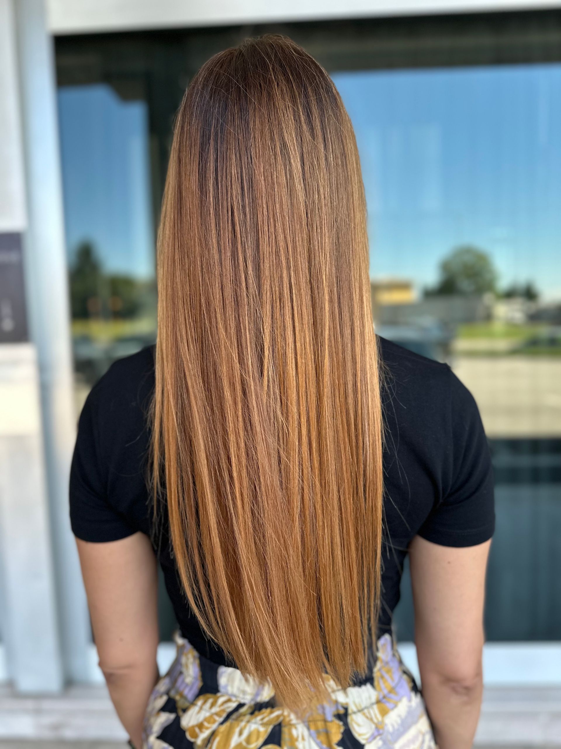 Una persona di spalle, con lunghi capelli castano dorato e ondulati, che indossa una camicia nera e una gonna a fantasia, all'aperto.