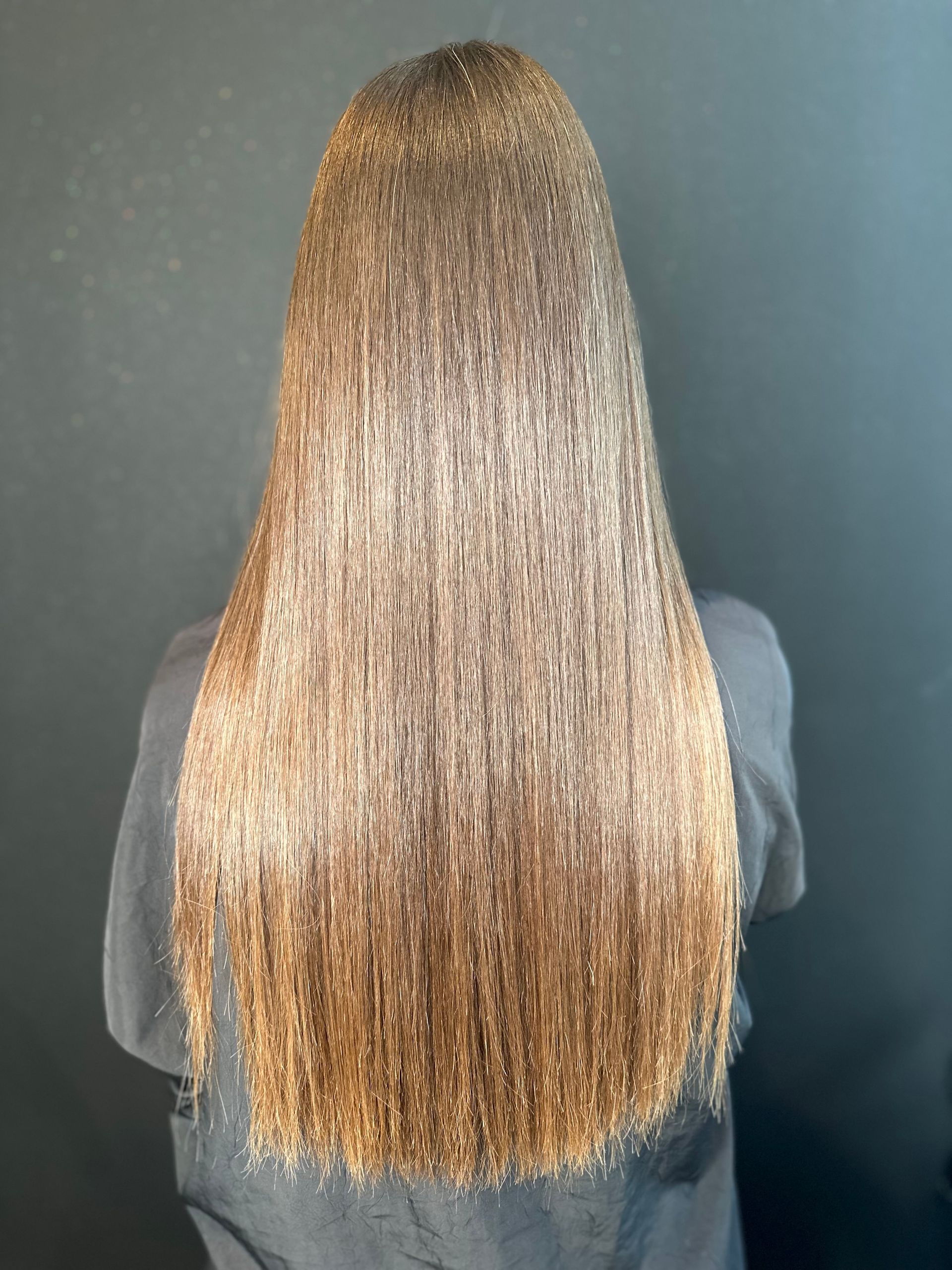 Vista posteriore di lunghi capelli castano chiaro, mossi e dalla texture particolare, che arrivano a metà schiena, su uno sfondo grigio scuro.
