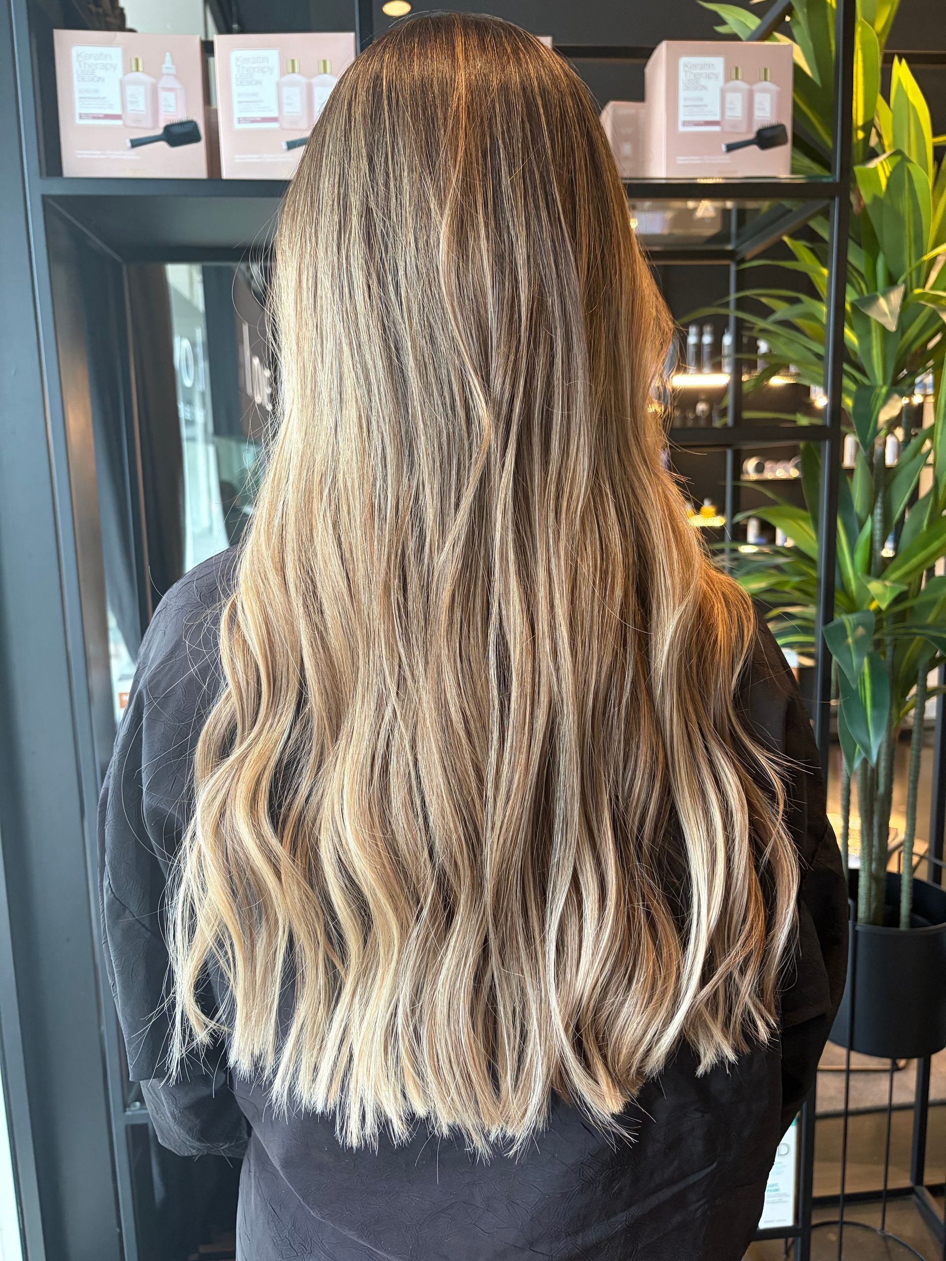 Una persona con lunghi capelli castano chiaro e dalla consistenza particolare, in un negozio con scaffali pieni di scatole di prodotti e una pianta verde sullo sfondo.