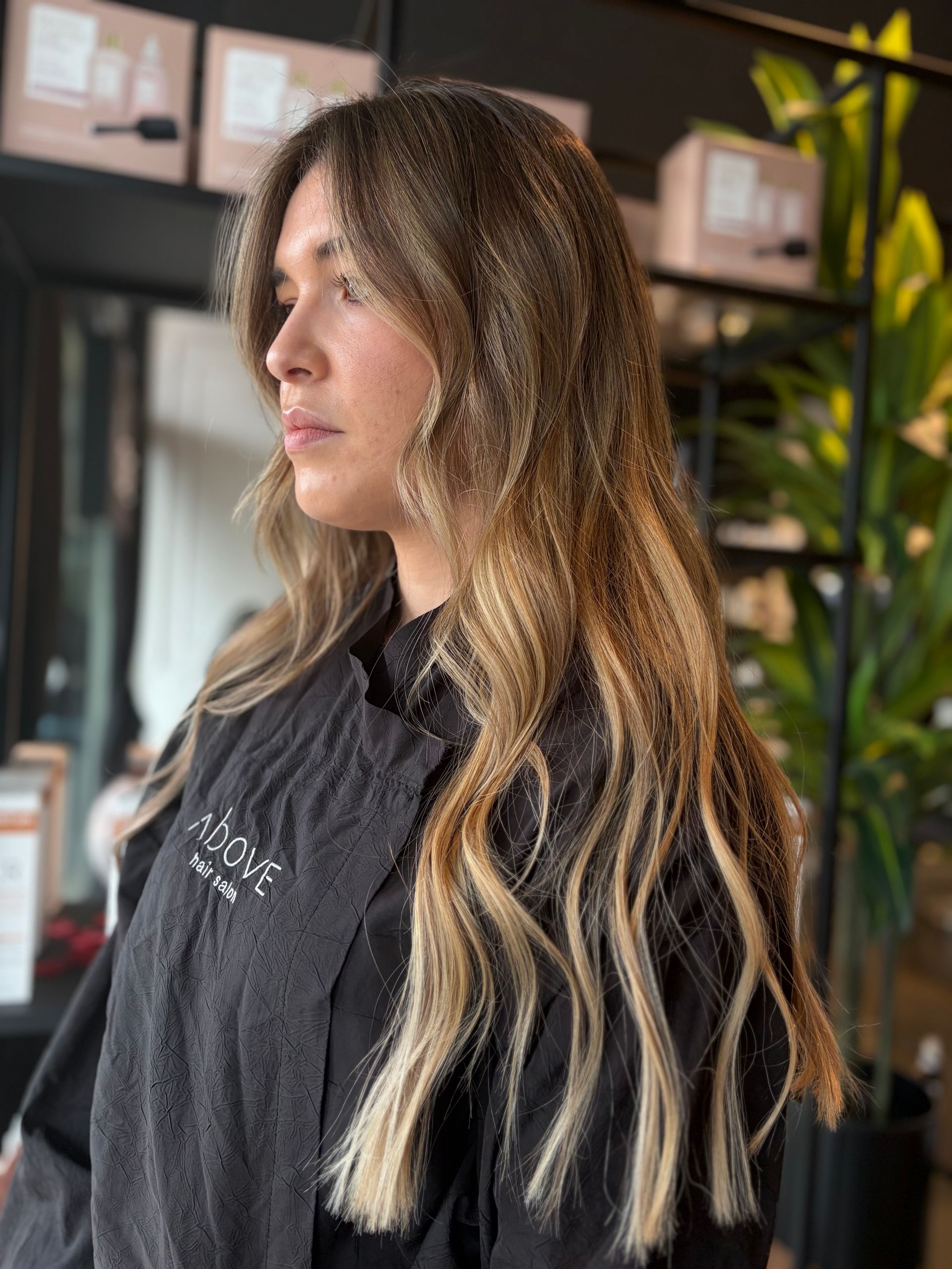 Una persona con lunghi capelli castani ondulati e con mèches, dalle punte bionde, in piedi in un salone con un mantello nero.