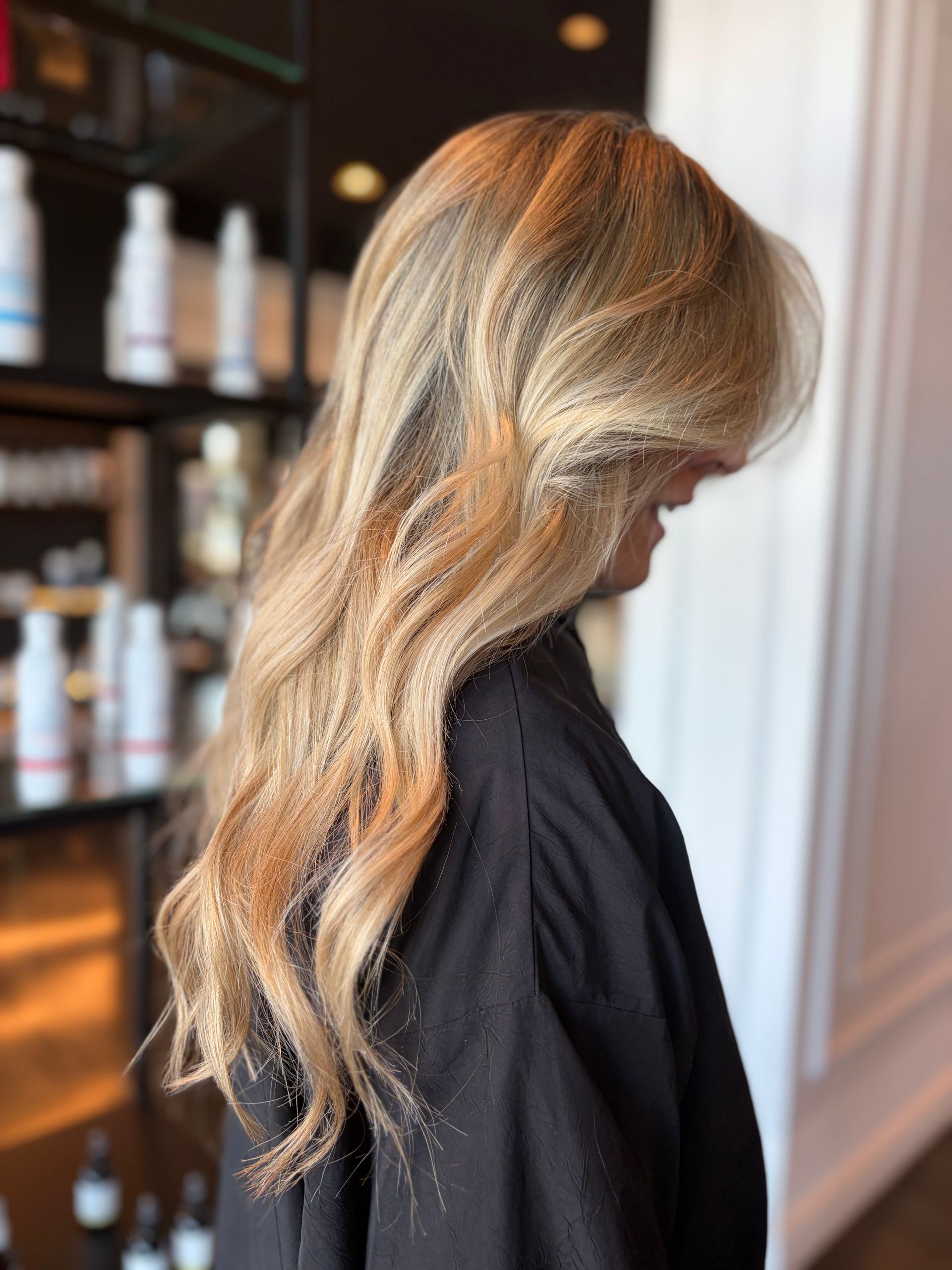 Una vista di profilo di lunghi capelli biondi ondulati con mèches in un salone di bellezza, con scaffali di prodotti sullo sfondo.