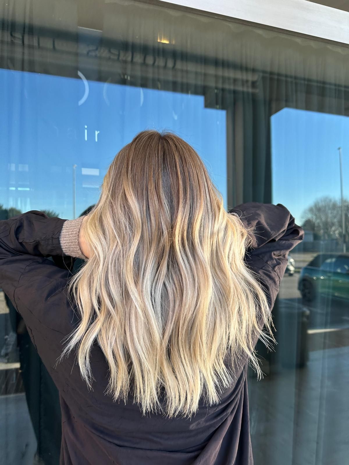 Una persona con capelli biondi ondulati e con mèches, vista di spalle, in piedi davanti a una vetrina.