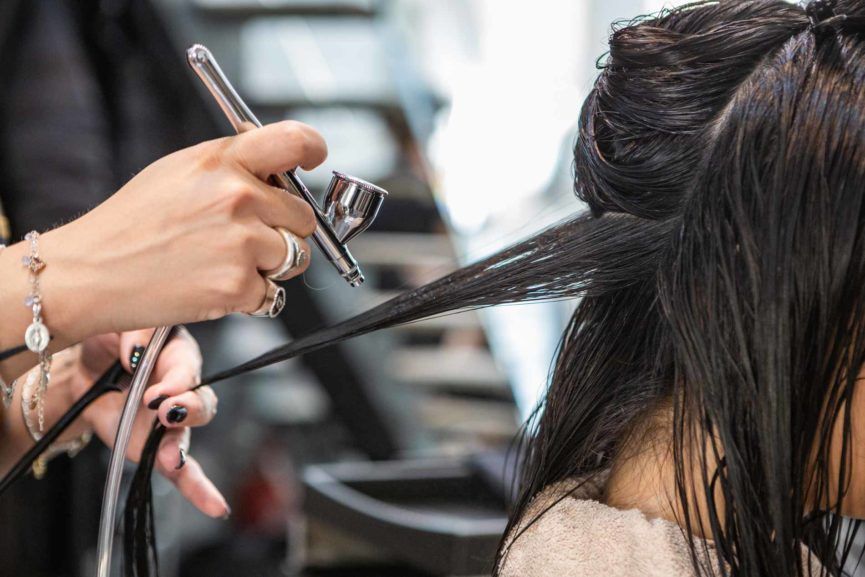 In un salone di bellezza, un parrucchiere utilizza un aerografo per applicare un trattamento per capelli su una sezione di capelli scuri e bagnati.