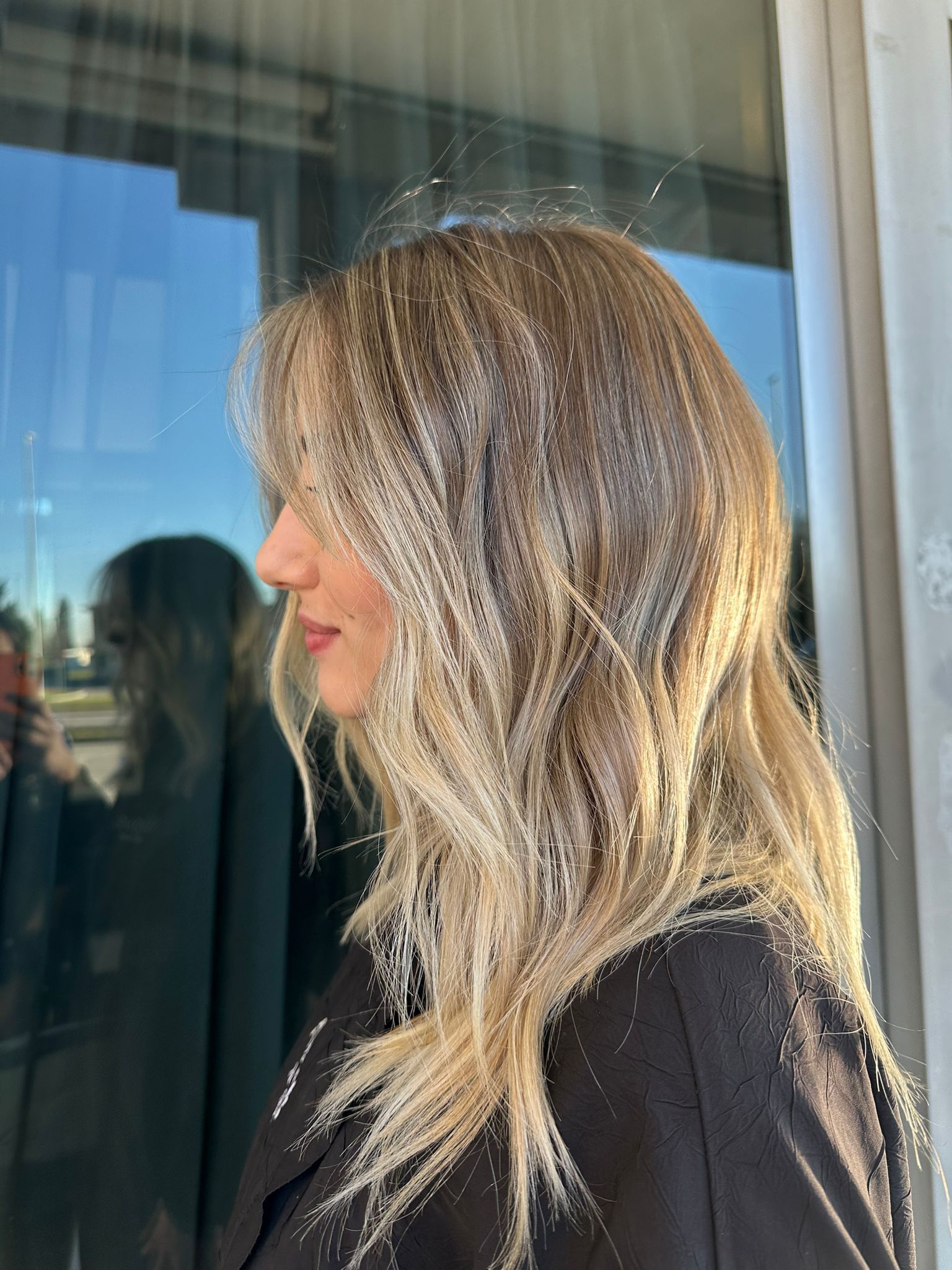 Profilo laterale di una persona con capelli mossi e biondi con mèches, in piedi all'aperto vicino a una finestra.