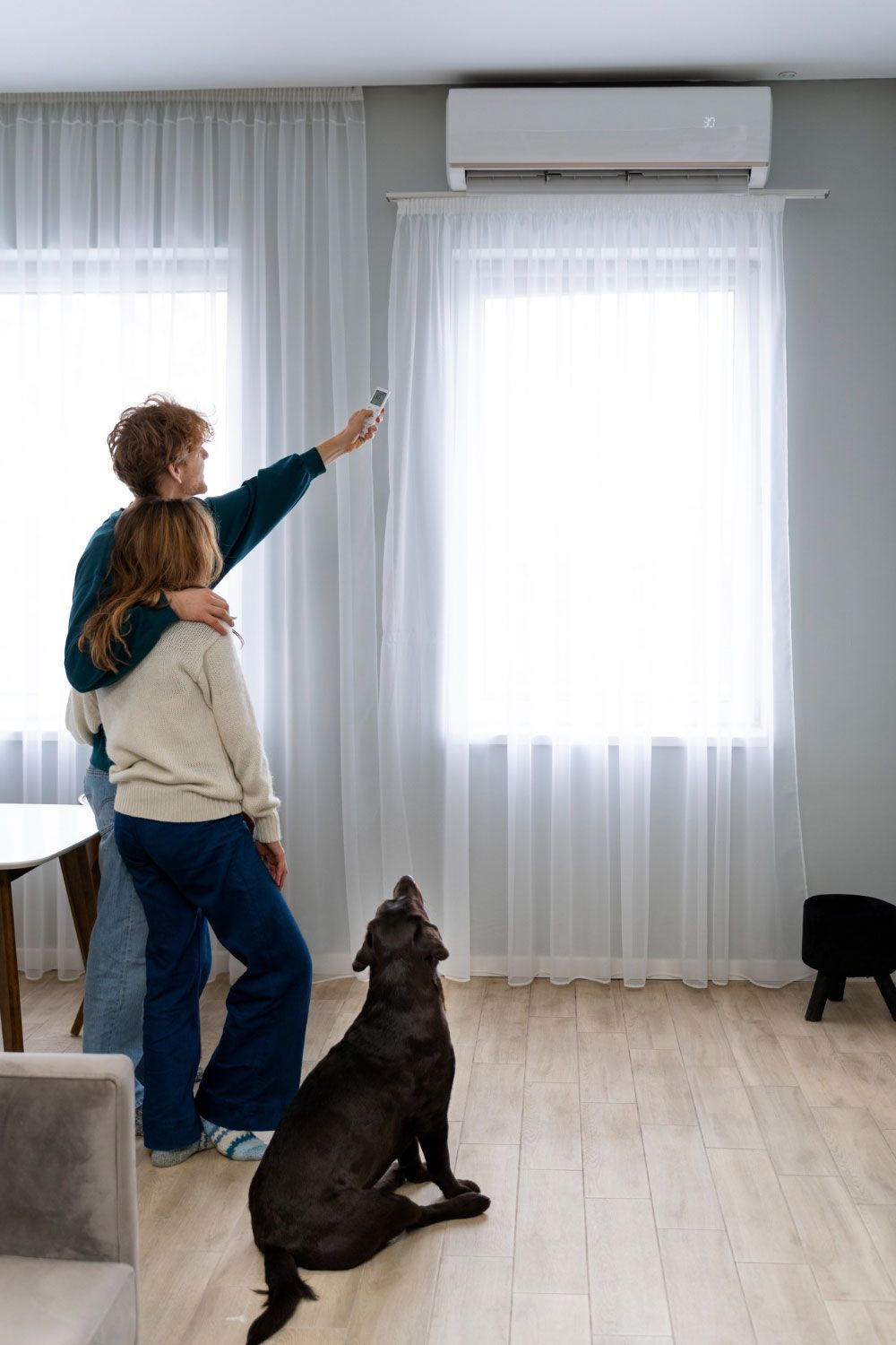 A man and a woman are standing in a living room with a dog and a remote control.