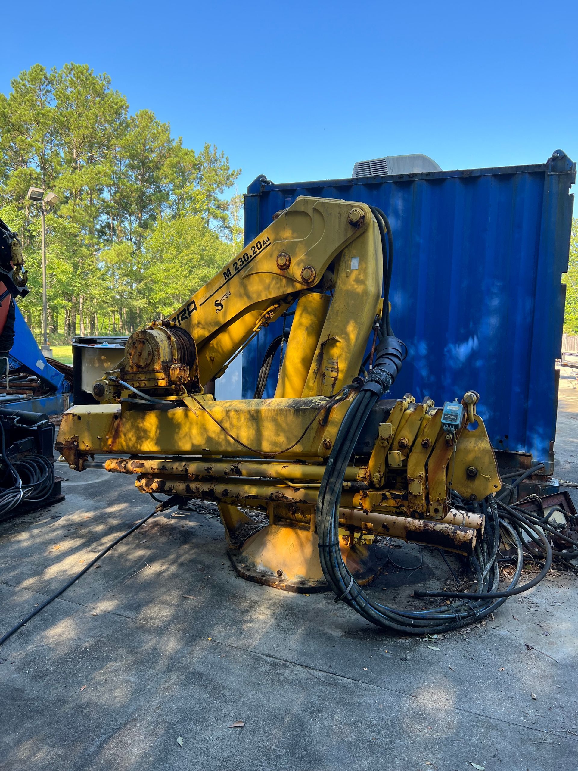Yellow industrial excavator arm and mechanism in front of a blue container on concrete.