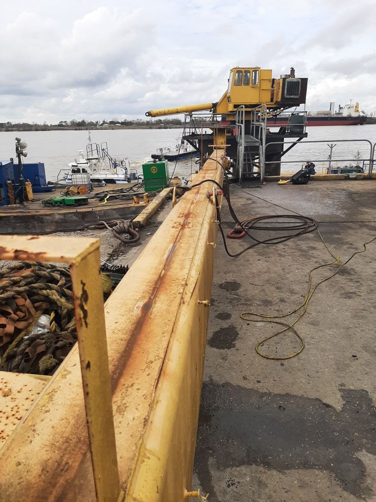 Yellow industrial arm extends from a dock with control cabin, pier, boats, water, and cloudy sky.