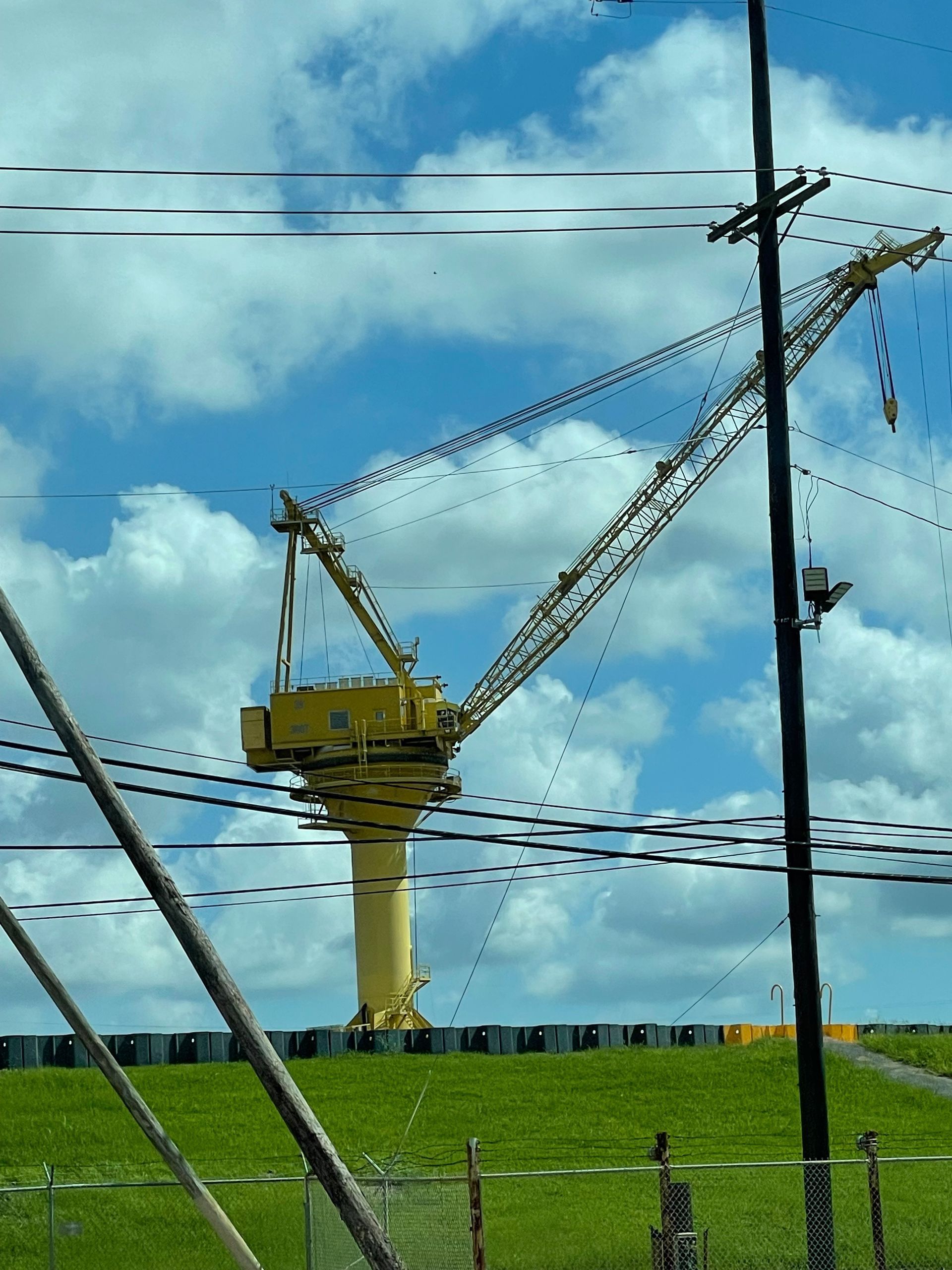 Yellow crane on a tall pillar, set against a cloudy blue sky and green landscape with power lines in the foreground.