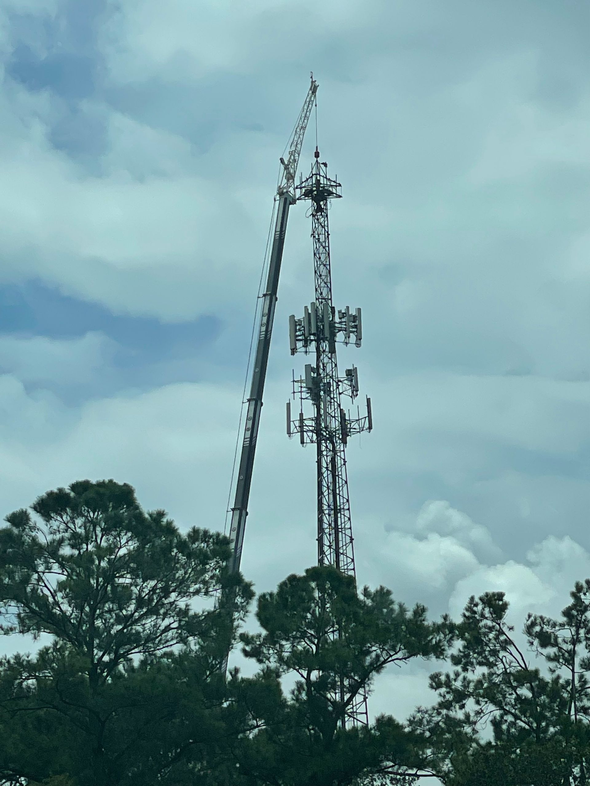 Cell tower with a crane working on the top section against a cloudy sky.