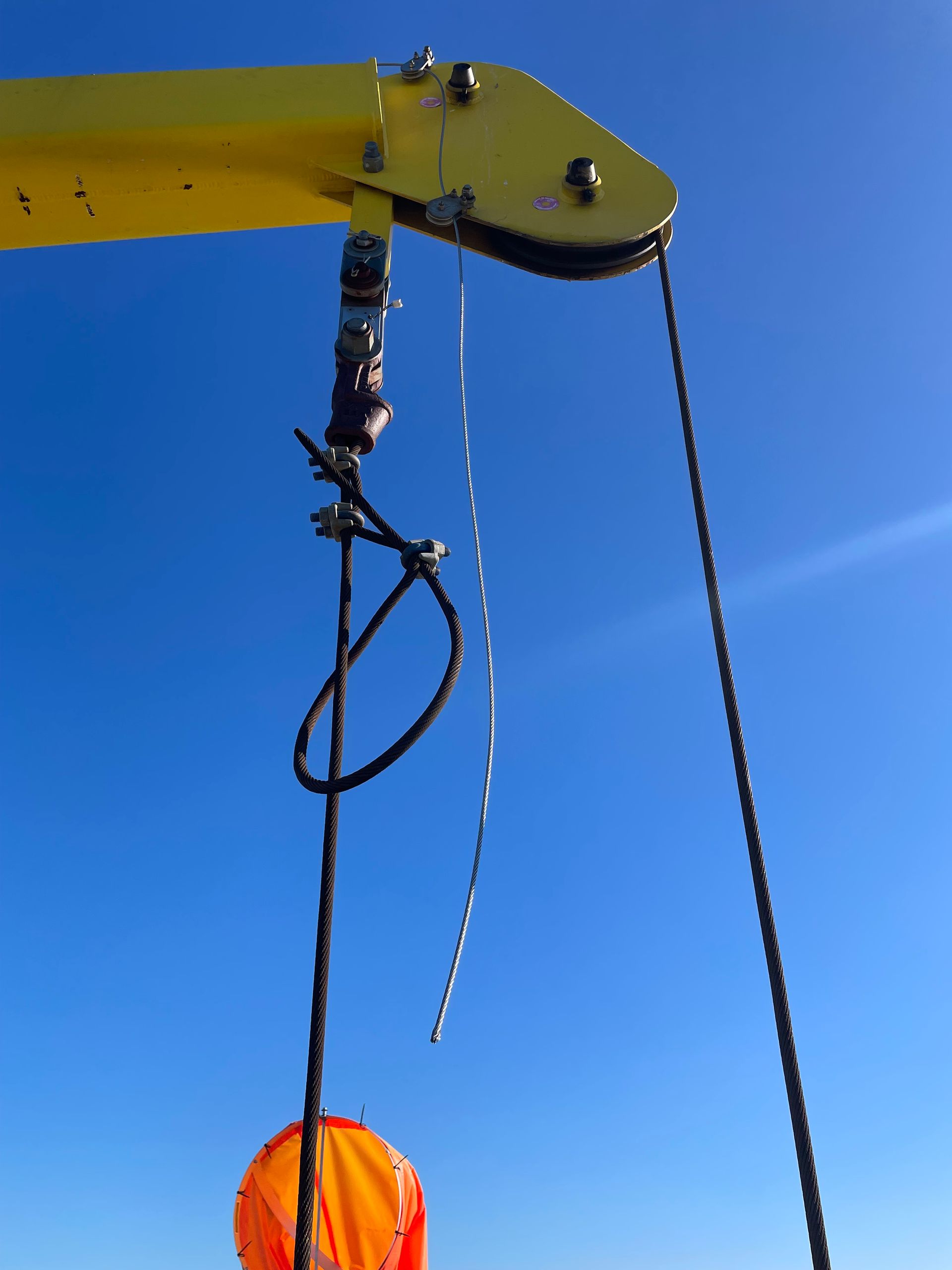 Yellow crane arm with pulley, cables, and orange buoy against a blue sky.